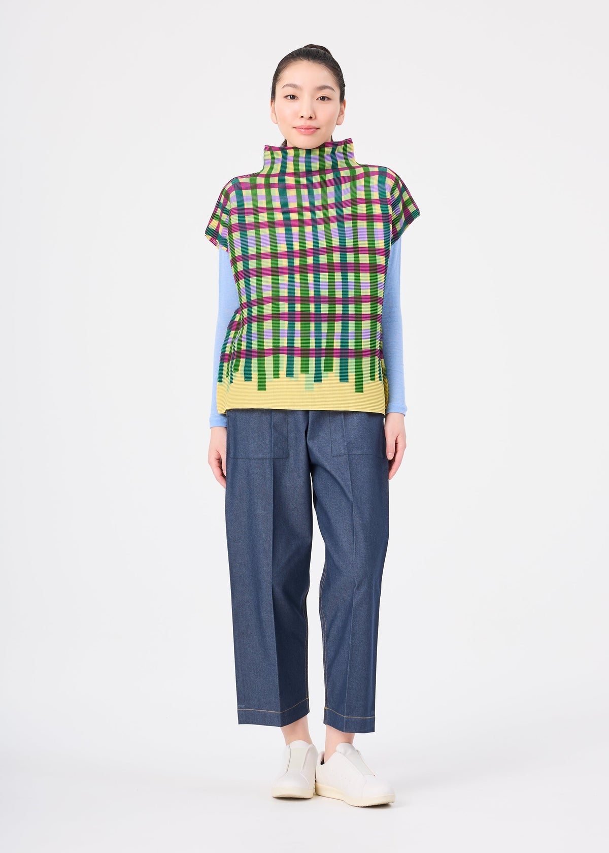 Zoomed view A person stands facing forward, wearing the TAPE CHECK top by me ISSEY MIYAKE over a light blue long-sleeve shirt, with loose dark blue pants and white shoes, against a plain white background.