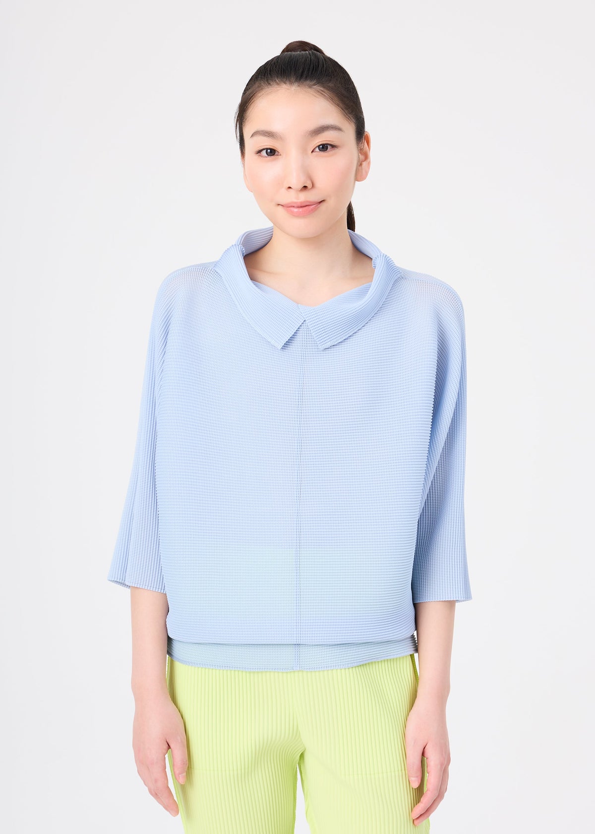 A woman with long dark hair in a ponytail wears the me ISSEY MIYAKE DOLMAN STRETCH PLEATS 1 top in light blue with three-quarter sleeves and a collar, paired with light green pants, against a plain white background. view 6 of 8