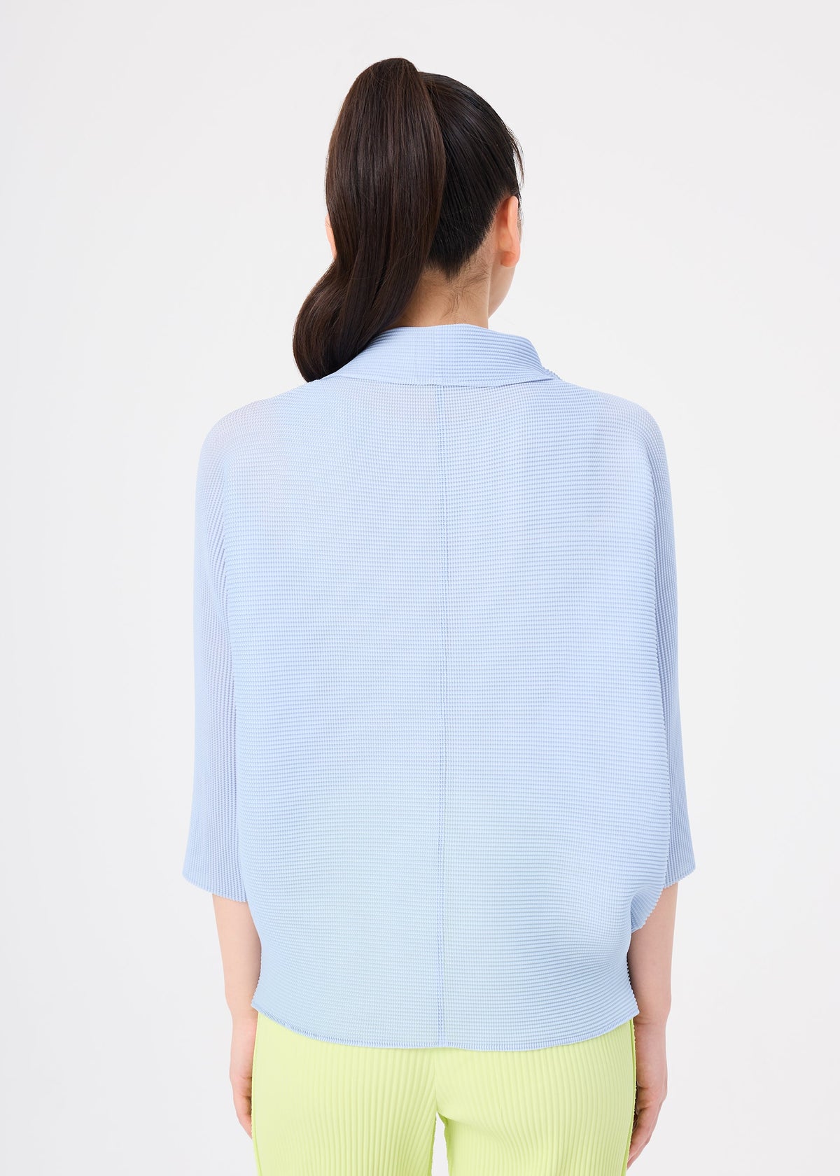 A person with long dark hair in a high ponytail is seen from the back, wearing the me ISSEY MIYAKE DOLMAN STRETCH PLEATS 1 light blue top and light yellow pants against a plain white background. view 7 of 8