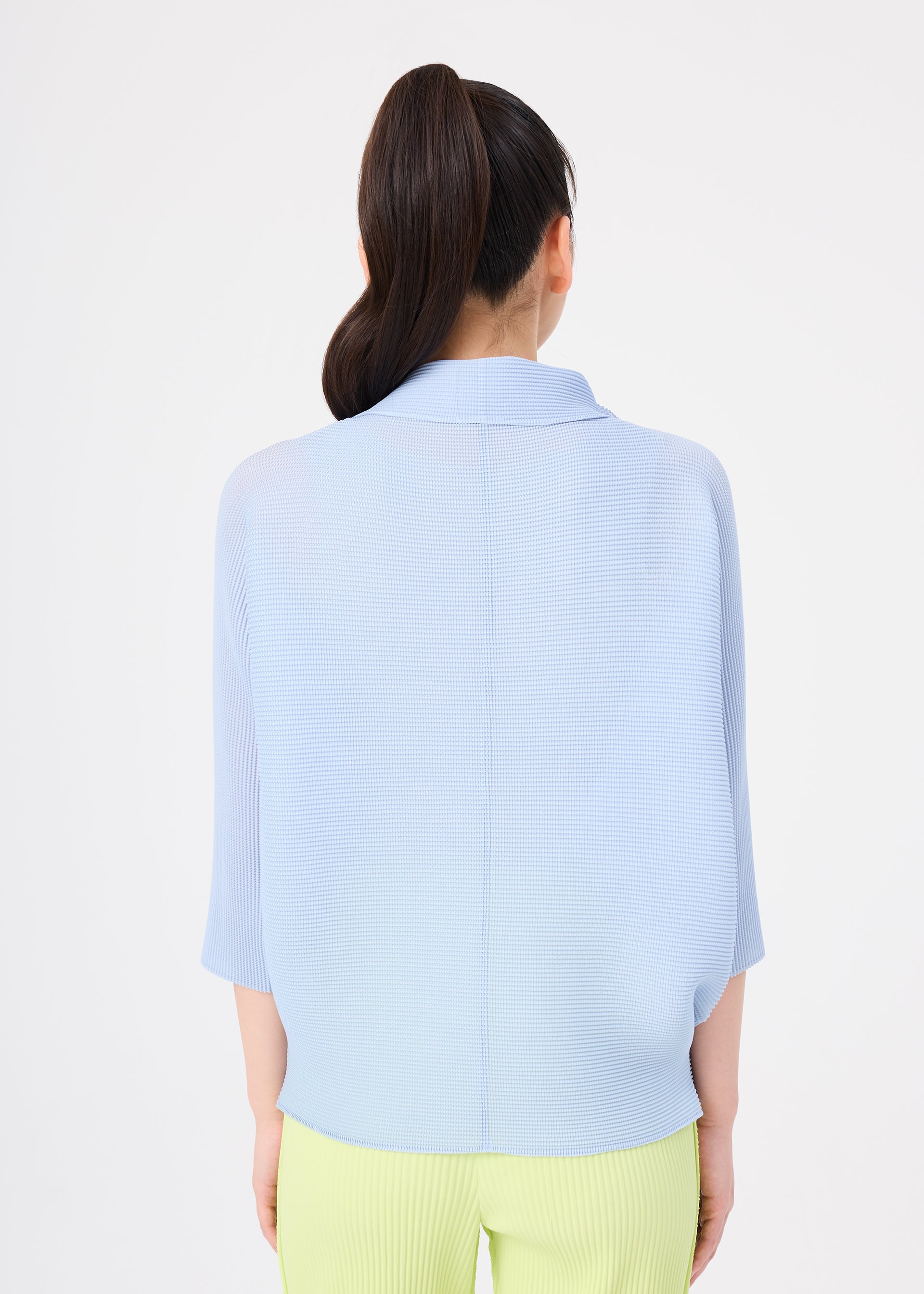 Zoomed view A person with long dark hair in a high ponytail is seen from the back, wearing the me ISSEY MIYAKE DOLMAN STRETCH PLEATS 1 light blue top and light yellow pants against a plain white background.