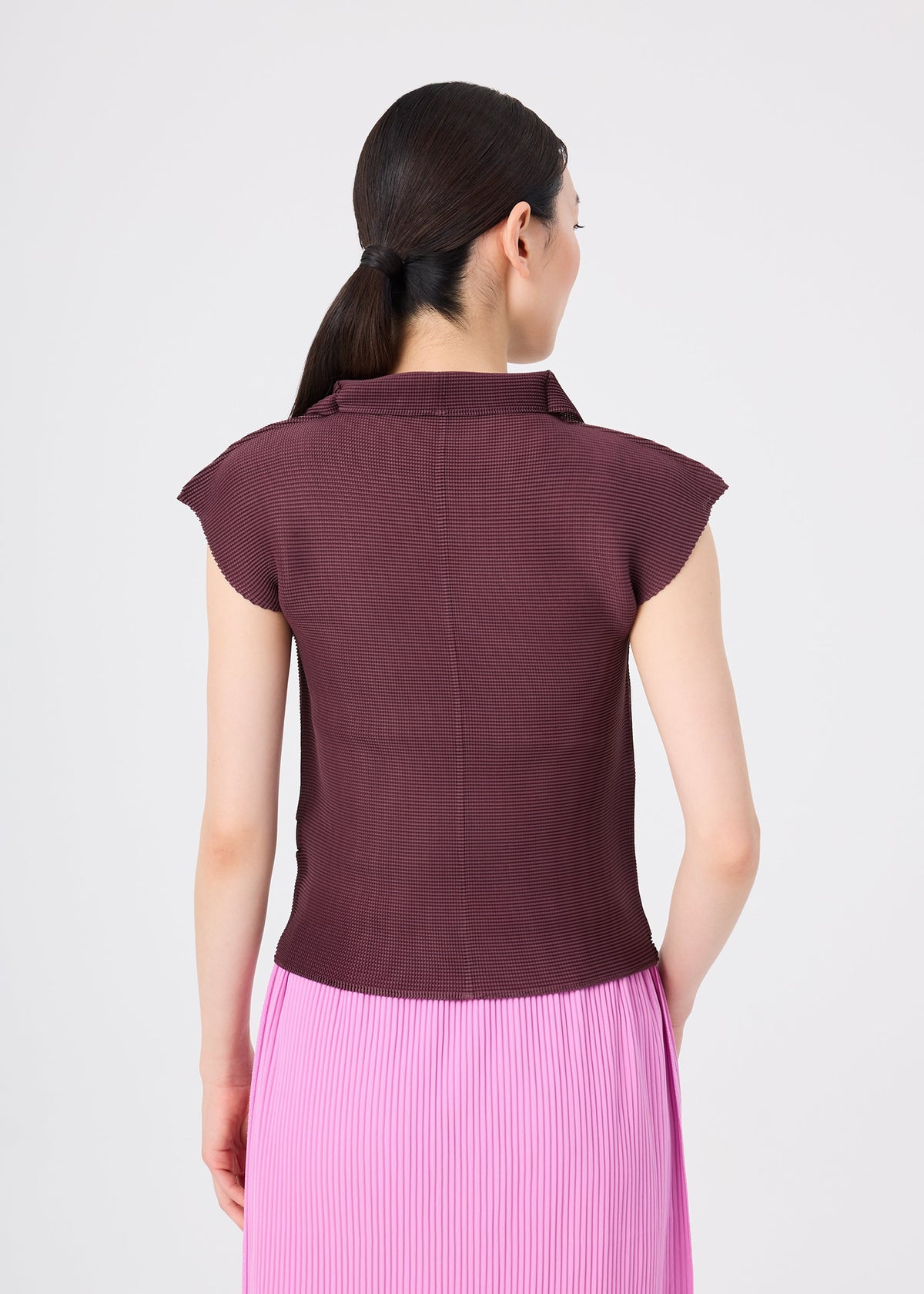 A woman with dark hair in a ponytail is shown from the back, wearing me ISSEY MIYAKE’s STRETCH PLEATS 2 top with French sleeves in maroon and a pleated pink skirt, set against a plain white background. view 6 of 7