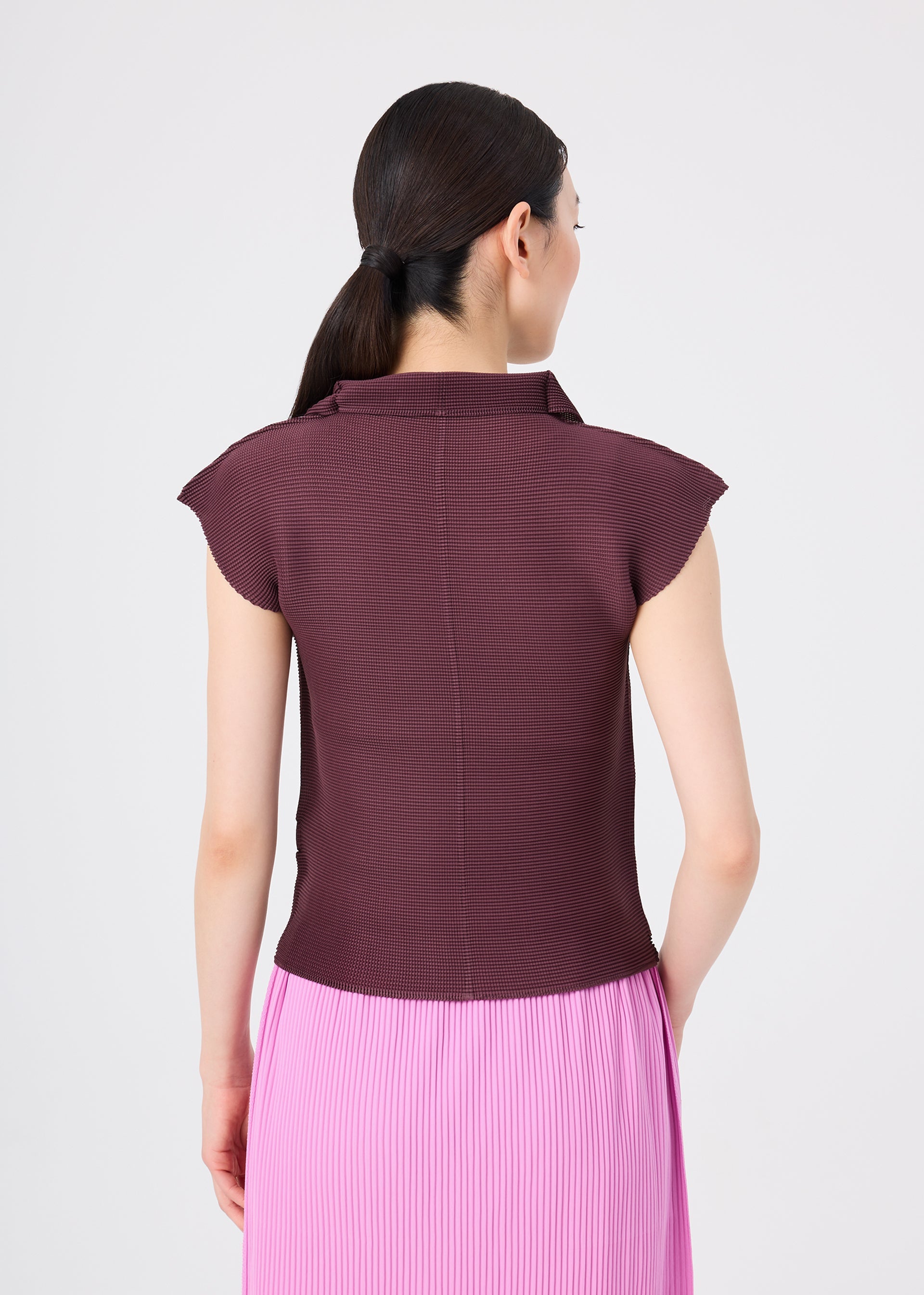 Zoomed view A woman with dark hair in a ponytail is shown from the back, wearing me ISSEY MIYAKE’s STRETCH PLEATS 2 top with French sleeves in maroon and a pleated pink skirt, set against a plain white background.