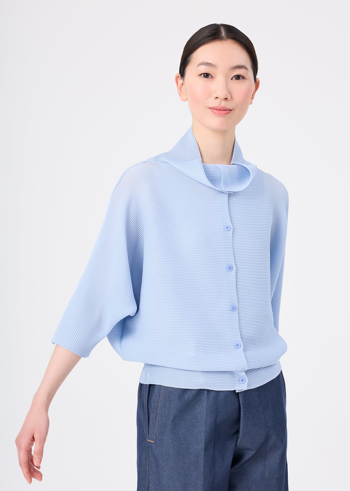 A woman with straight dark hair wears the me ISSEY MIYAKE DOLMAN STRETCH PLEATS 1, a light blue textured button-up with three-quarter sleeves, paired with dark blue jeans against a plain white background. view 5 of 7