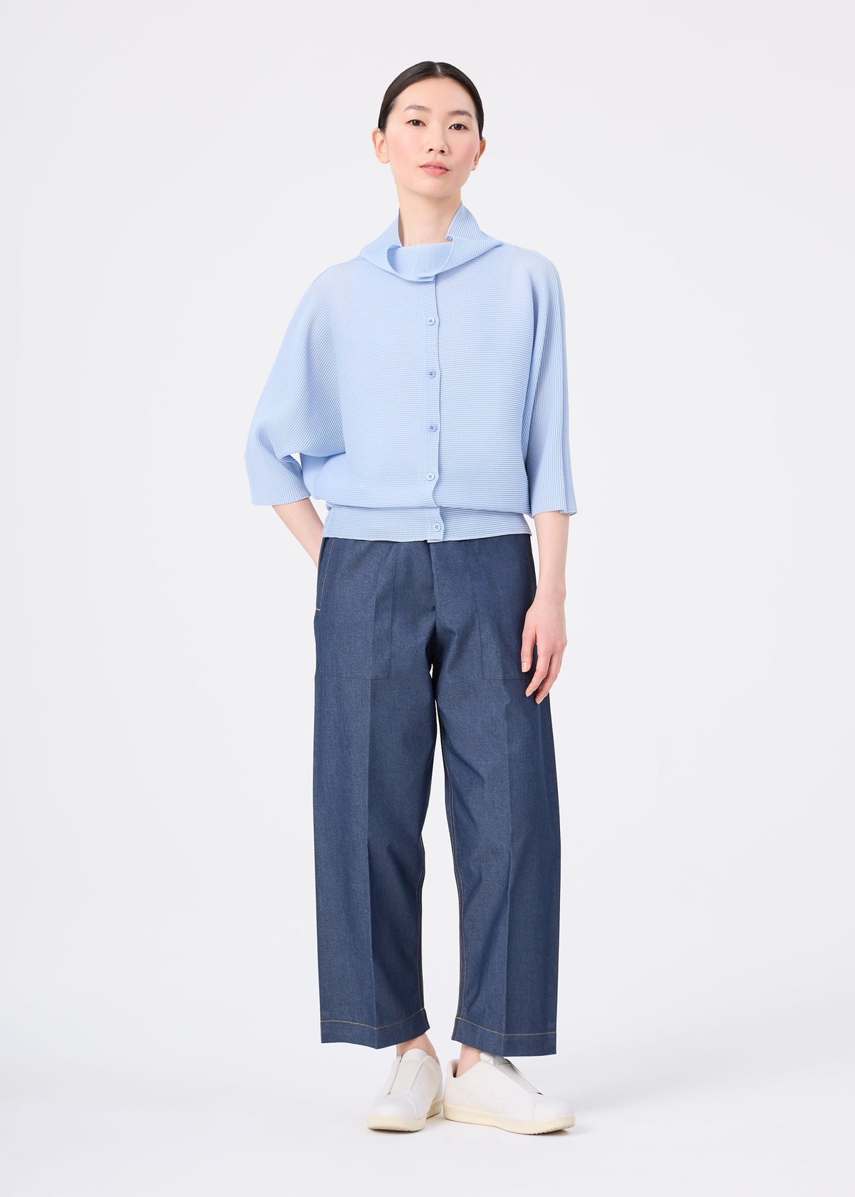 A woman with dark hair pulled back wears the me ISSEY MIYAKE DOLMAN STRETCH PLEATS 1 light blue three-quarter sleeve button-up top, wide-leg dark blue pants, and white slip-on shoes against a plain white background. view 7 of 7