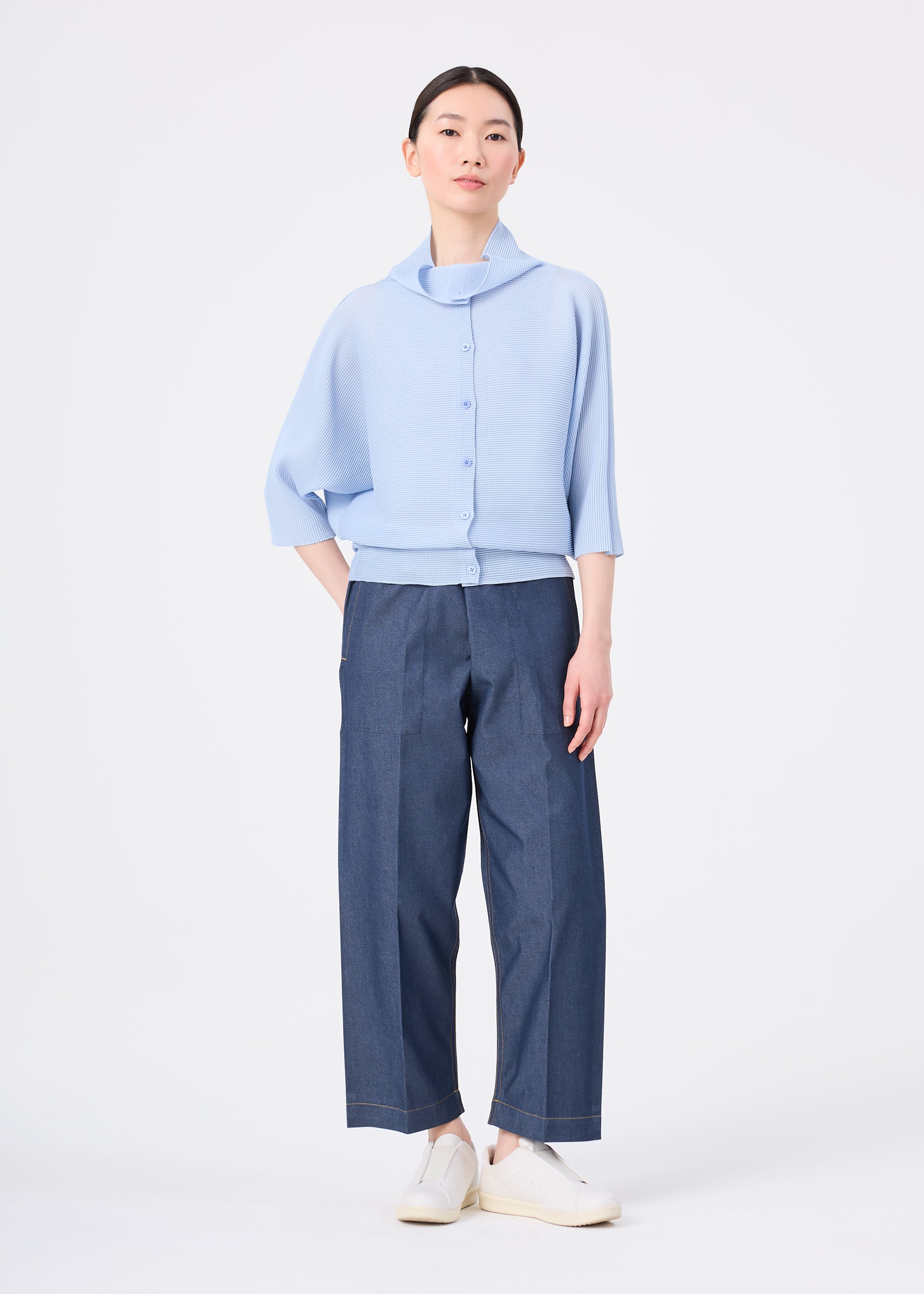 Zoomed view A woman with dark hair pulled back wears the me ISSEY MIYAKE DOLMAN STRETCH PLEATS 1 light blue three-quarter sleeve button-up top, wide-leg dark blue pants, and white slip-on shoes against a plain white background.