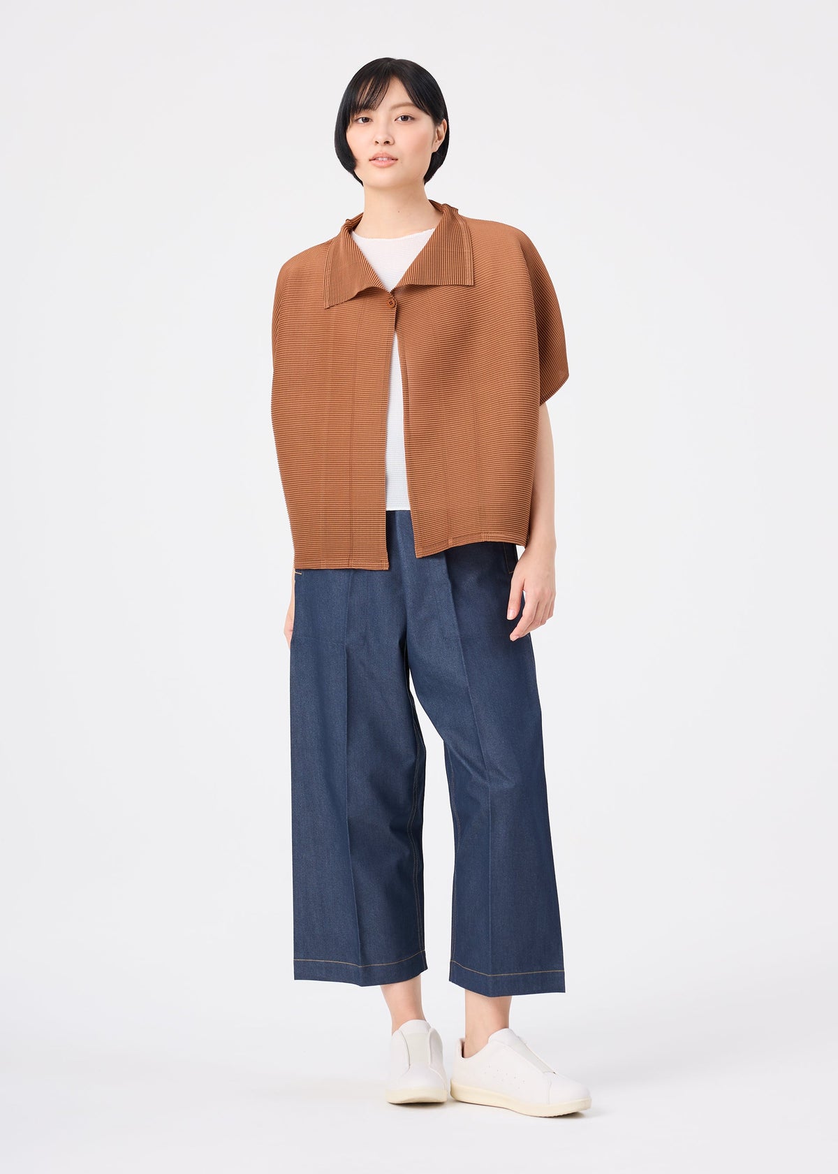 A person stands against a white background wearing the me ISSEY MIYAKE SQUARE STRETCH PLEATS relaxed fit brown short-sleeve jacket over a white shirt, paired with wide-leg blue denim pants and white slip-on shoes. view 9 of 10