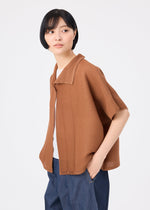 A person with short black hair wears the me ISSEY MIYAKE SQUARE STRETCH PLEATS, a brown, relaxed-fit short-sleeved cardigan, over a white shirt and blue pants, standing against a plain white background and looking to the side.