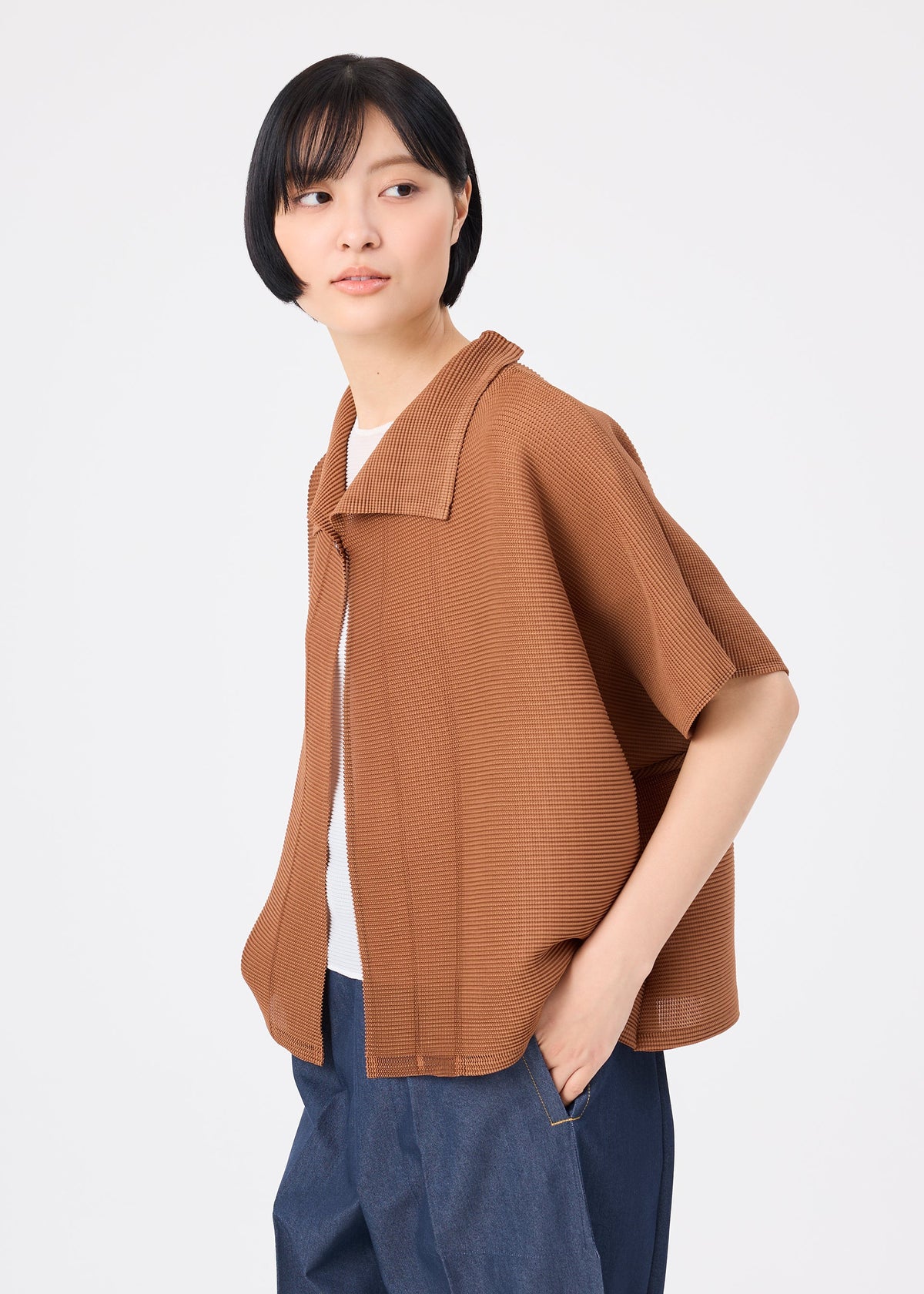 Zoomed view A person with short black hair wears the me ISSEY MIYAKE SQUARE STRETCH PLEATS, a brown, relaxed-fit short-sleeved cardigan, over a white shirt and blue pants, standing against a plain white background and looking to the side.