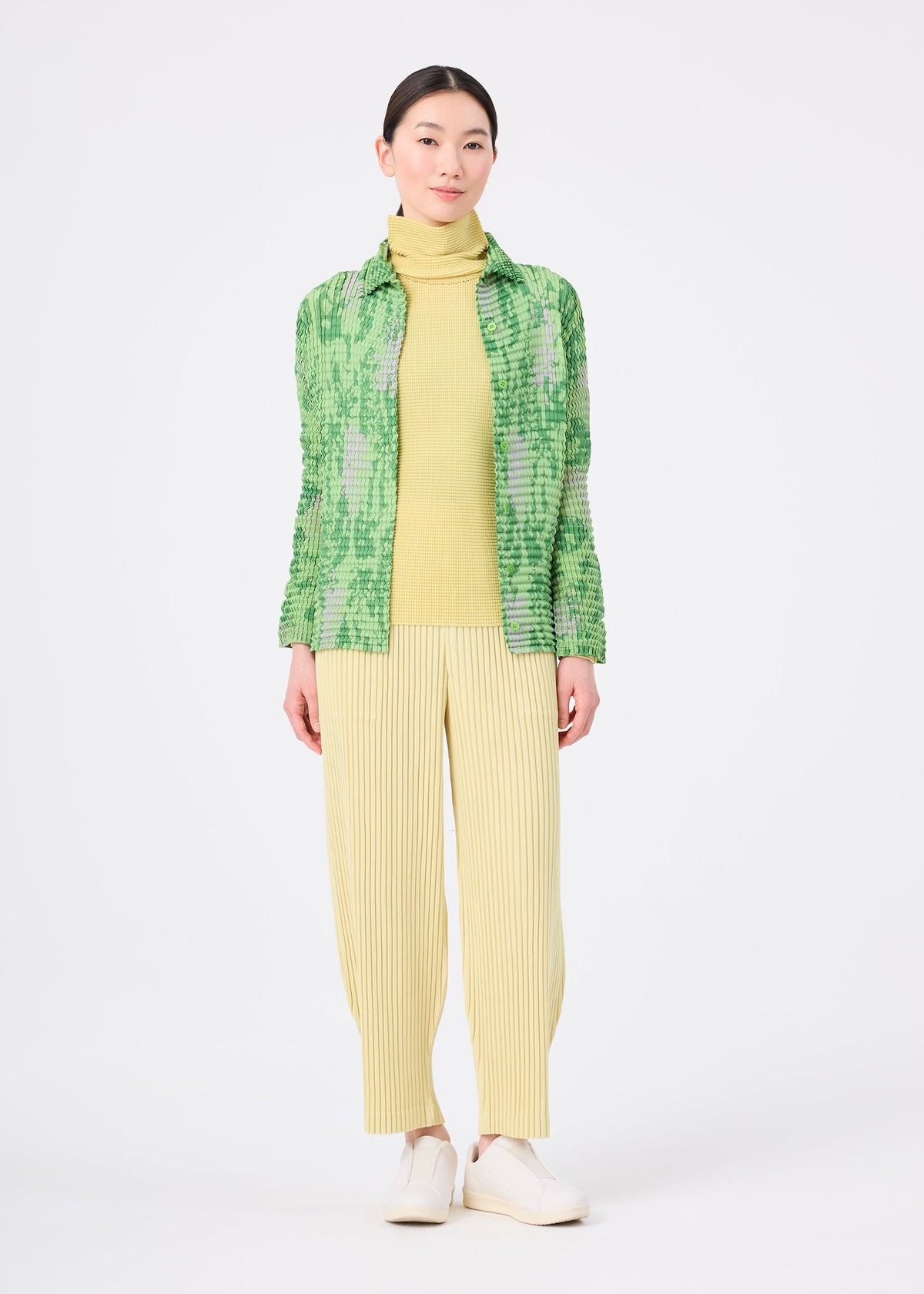Zoomed view A woman faces forward wearing the LEAF PLEATS SHIRT by me ISSEY MIYAKE over a yellow turtleneck, yellow pleated pants, and white shoes against a plain white background.