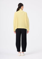A woman stands facing away in the me ISSEY MIYAKE CORDUROY PLEATS, a loose yellow ribbed sweater, paired with black wide pants and white shoes against a plain white background.