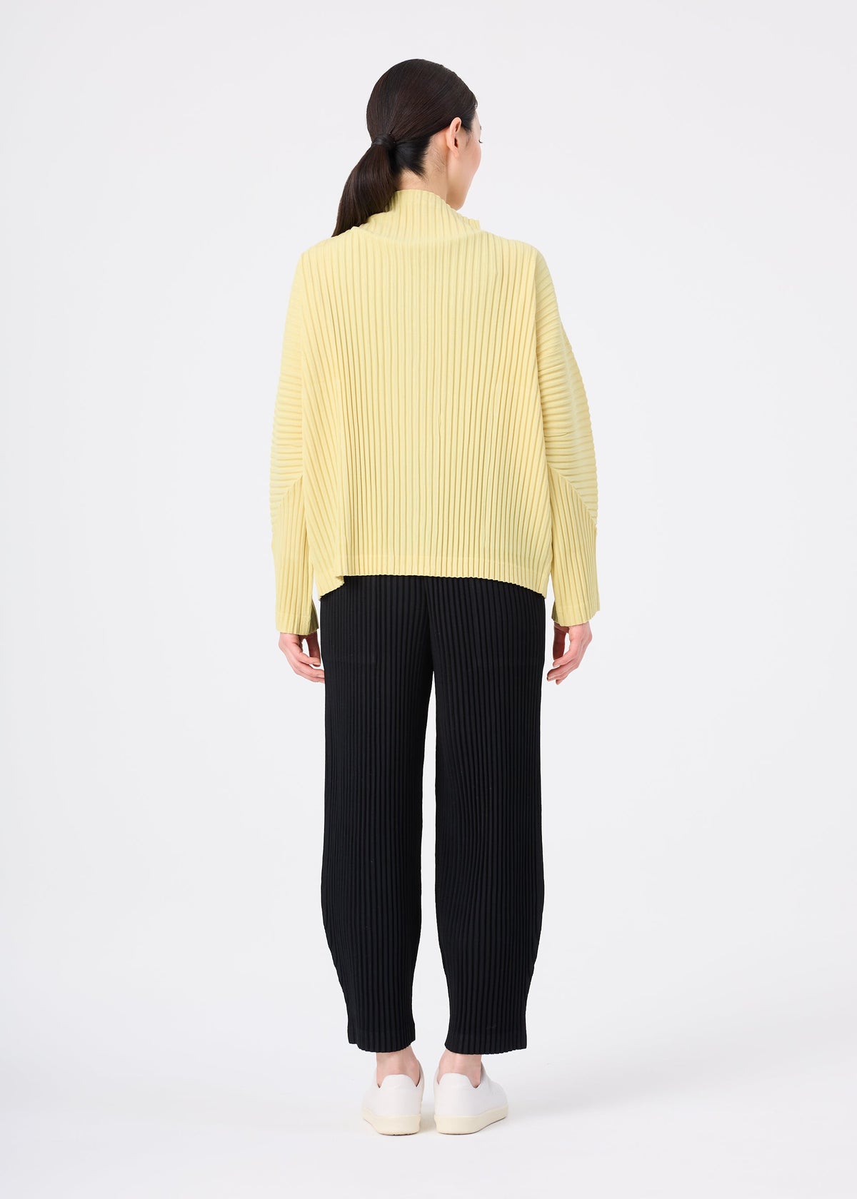 Zoomed view A woman stands facing away in the me ISSEY MIYAKE CORDUROY PLEATS, a loose yellow ribbed sweater, paired with black wide pants and white shoes against a plain white background.