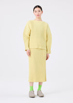 A person stands against a white background wearing the me ISSEY MIYAKE CORDUROY PLEATS skirt, paired with a loose, ribbed, light yellow top, green socks, and beige shoes.