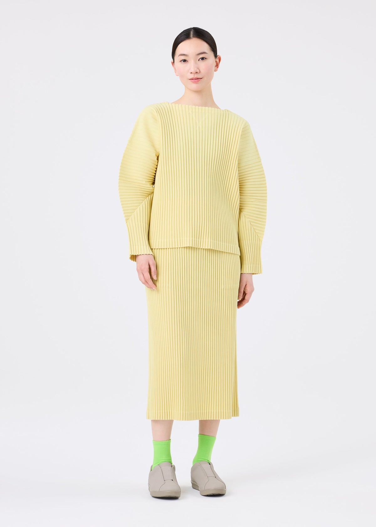 Zoomed view A person stands against a white background wearing the me ISSEY MIYAKE CORDUROY PLEATS skirt, paired with a loose, ribbed, light yellow top, green socks, and beige shoes.
