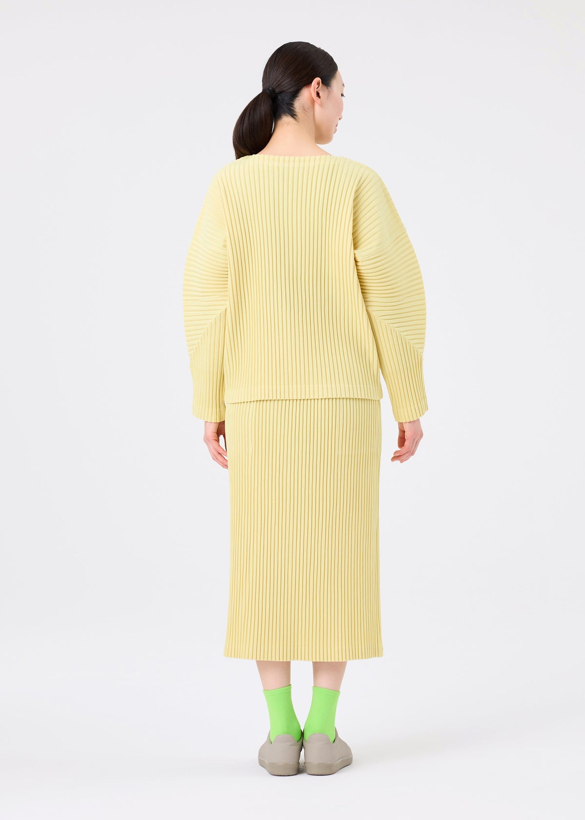 A person stands facing away in me ISSEY MIYAKE’s CORDUROY PLEATS, a ribbed light yellow sweater and soft brushed skirt, with dark hair tied back, green socks, and beige shoes against a plain white background. view 7 of 8