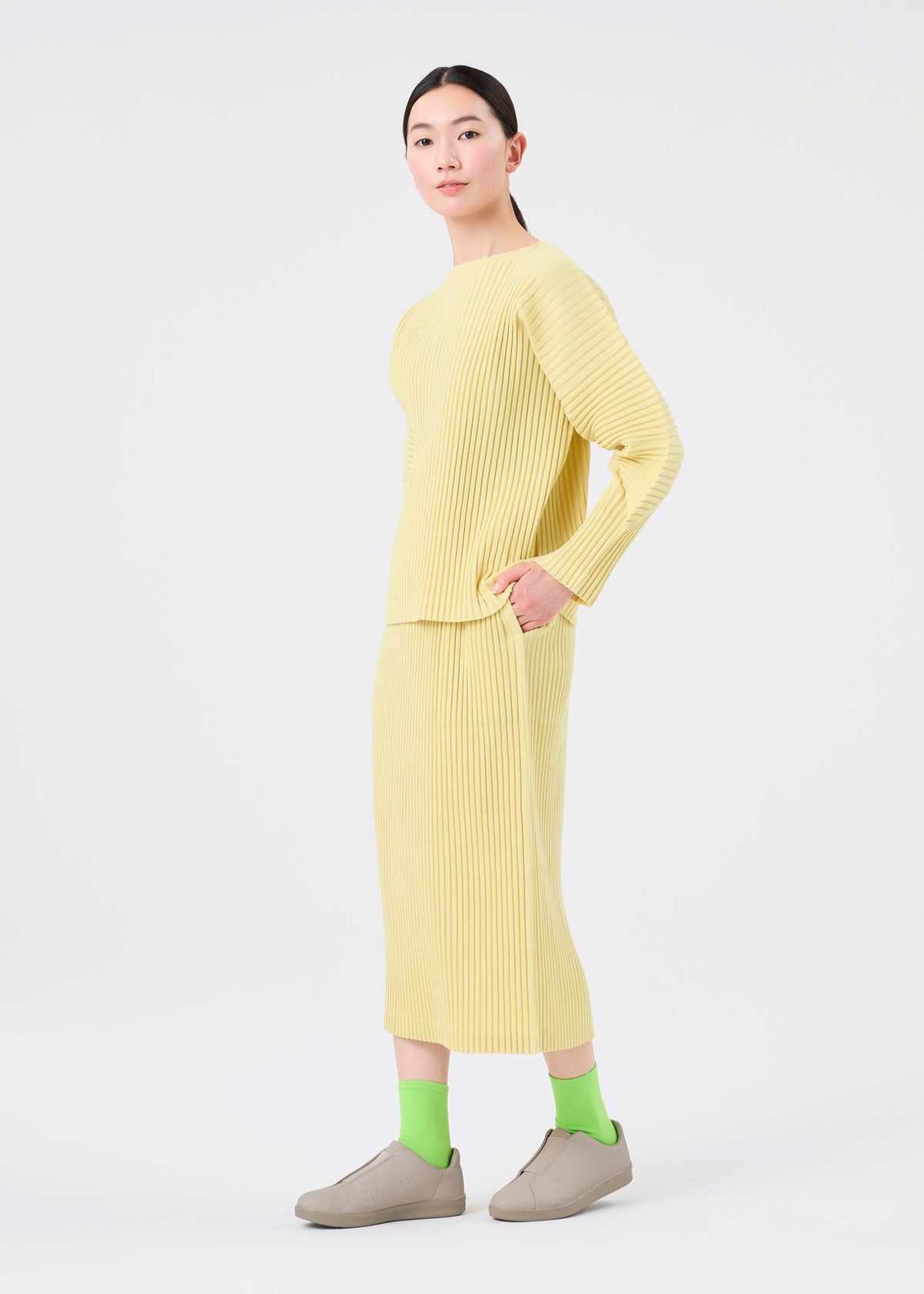A person stands against a white background wearing the me ISSEY MIYAKE CORDUROY PLEATS skirt with pockets and matching top, bright green socks, beige slip-on shoes, and has straight dark hair pulled back. view 8 of 8