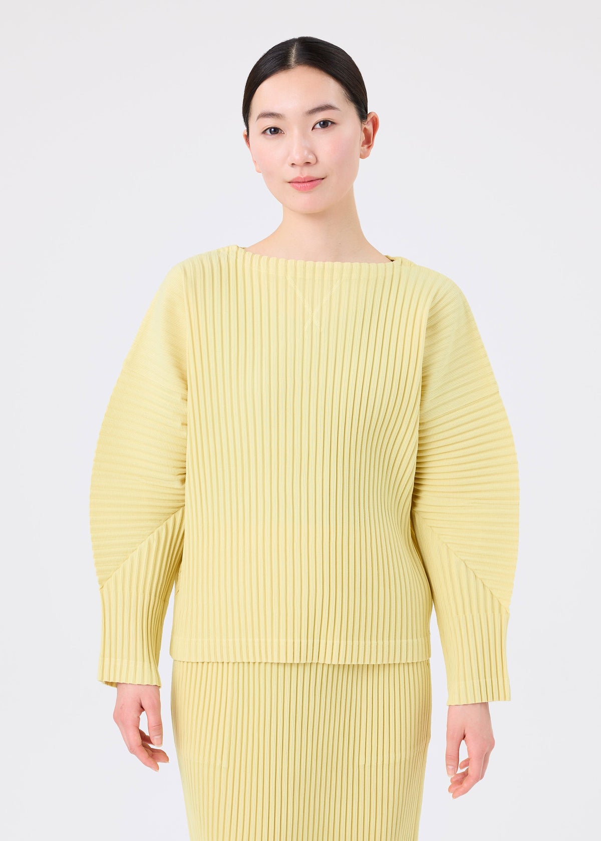 Zoomed view A person with dark hair in a low bun wears the light yellow CORDUROY PLEATS set by me ISSEY MIYAKE, featuring a pleated skirt and matching long-sleeved top, standing against a plain white background.