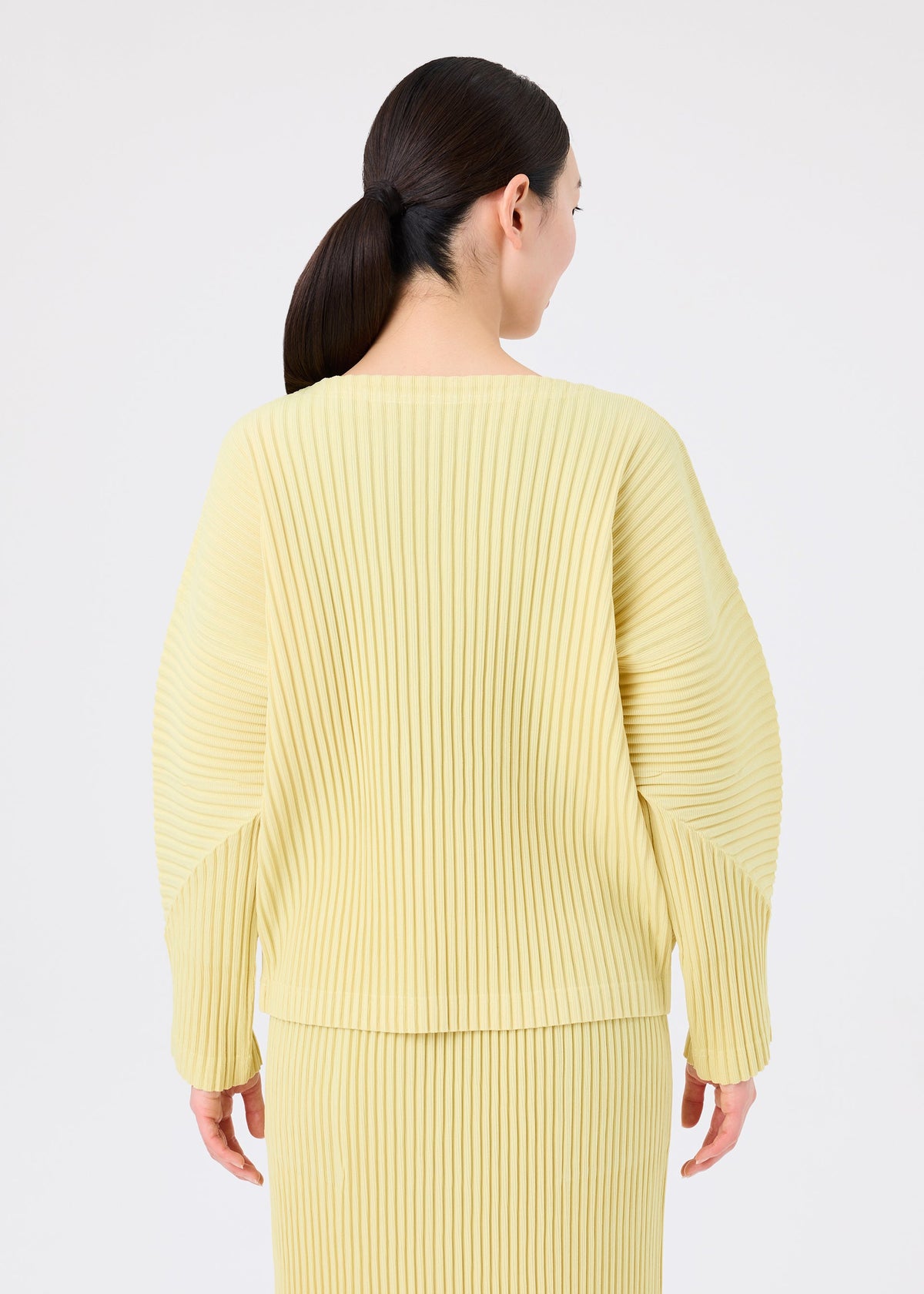 Zoomed view A woman with dark hair in a low ponytail is shown from the back wearing the CORDUROY PLEATS set by me ISSEY MIYAKE against a plain light background.
