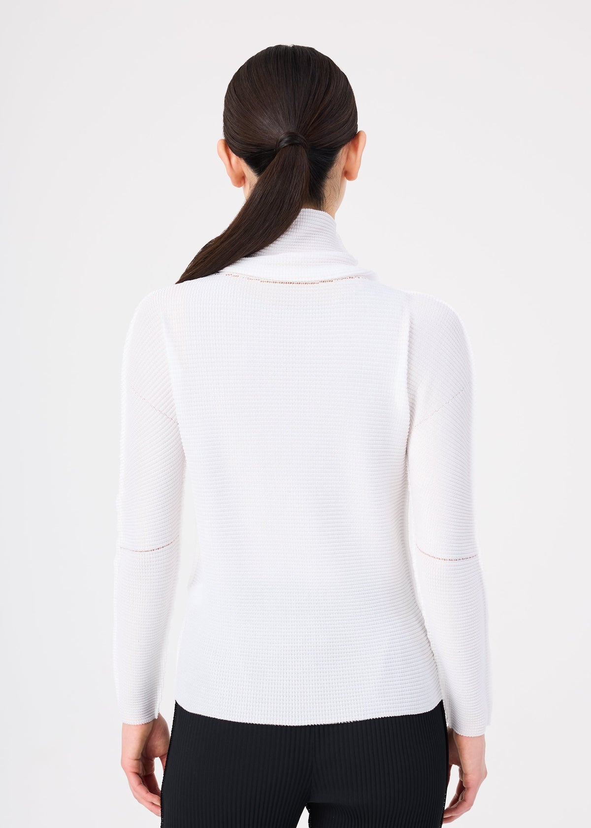 A woman is seen from behind, wearing the me ISSEY MIYAKE KNIT STRETCH PLEATS ribbed white long-sleeved cowl neck sweater with black pants. Her dark hair is styled in a sleek, low ponytail, set against a plain white background. view 4 of 5