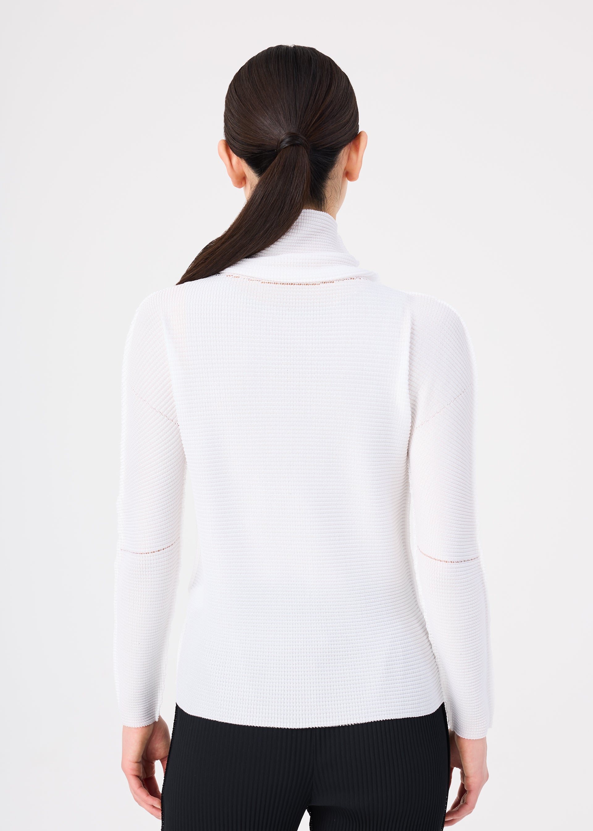 Zoomed view A woman is seen from behind, wearing the me ISSEY MIYAKE KNIT STRETCH PLEATS ribbed white long-sleeved cowl neck sweater with black pants. Her dark hair is styled in a sleek, low ponytail, set against a plain white background.