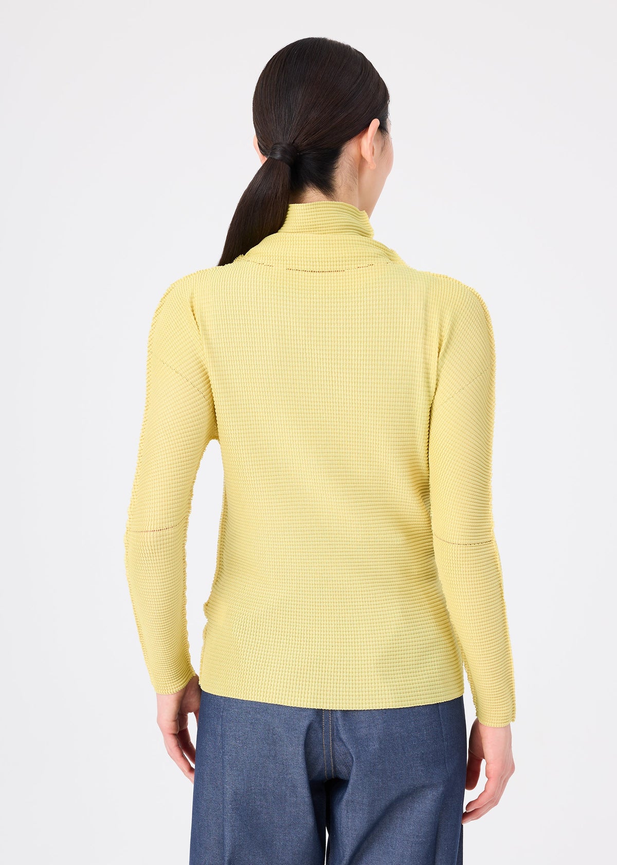 A person with long dark hair in a low ponytail wears the KNIT STRETCH PLEATS 1 top from me ISSEY MIYAKE and blue pants, facing away from the camera against a white background. view 6 of 7