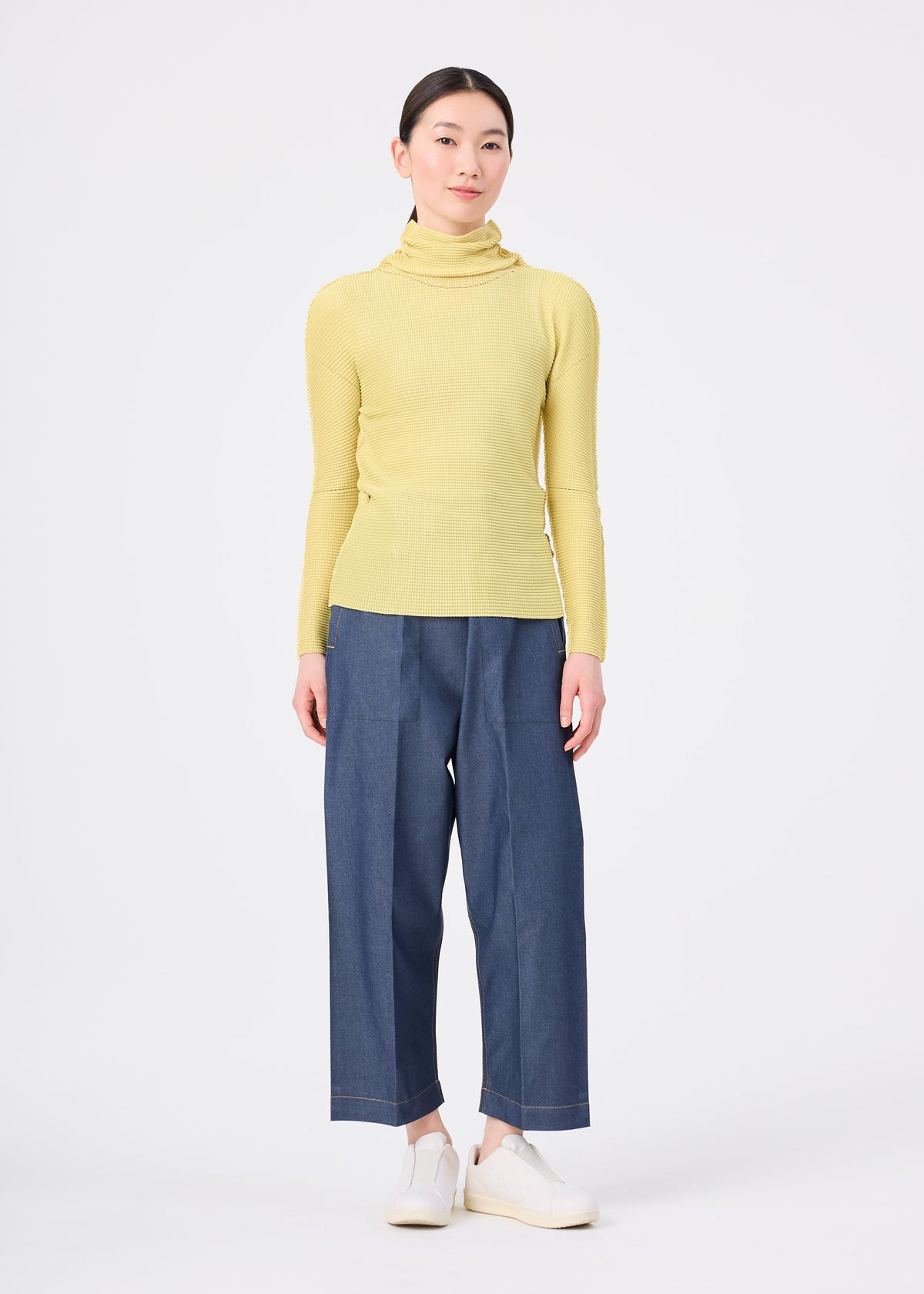 Zoomed view A person stands facing forward, wearing the KNIT STRETCH PLEATS 1 top by me ISSEY MIYAKE with blue wide-leg pants and white shoes, against a plain white background.
