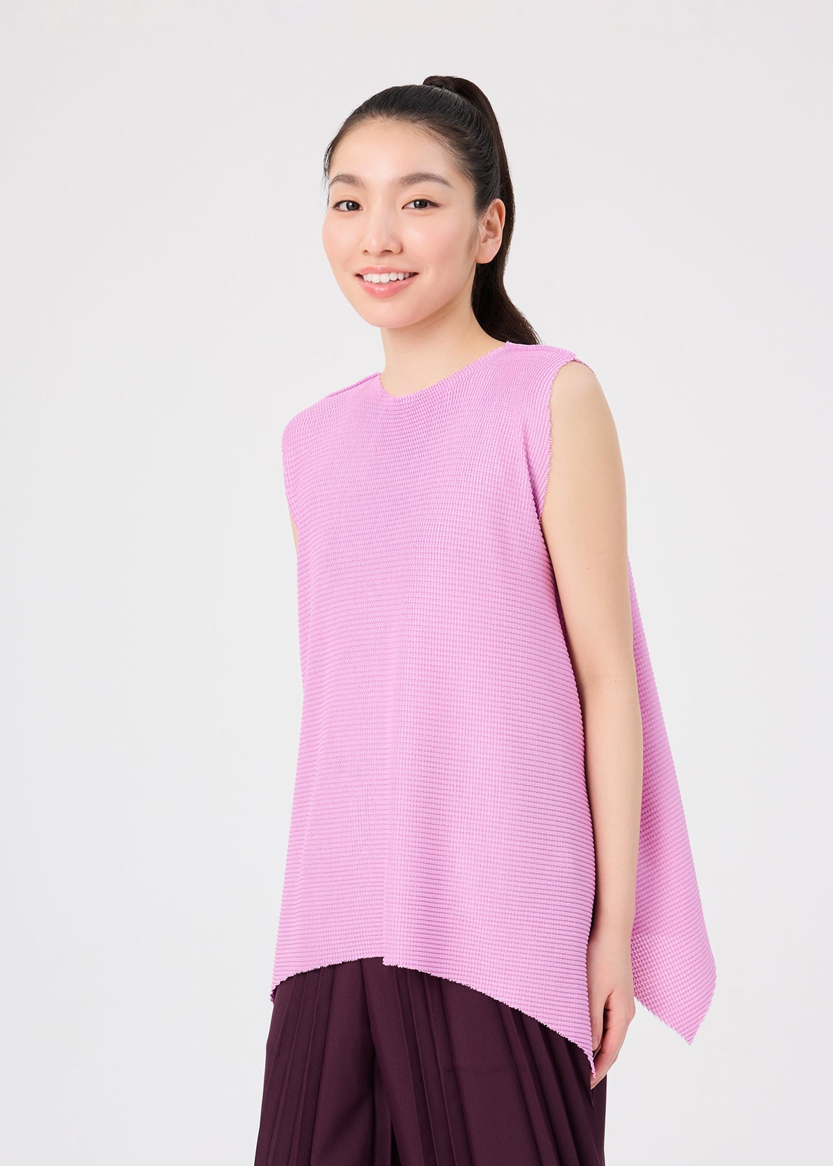 A woman with long dark hair in a ponytail wears the me ISSEY MIYAKE TOWER KNIT STRETCH PLEATS pants, paired with a sleeveless, soft-textured knit top featuring an asymmetric hem. She smiles as she stands against a plain light background. view 7 of 9