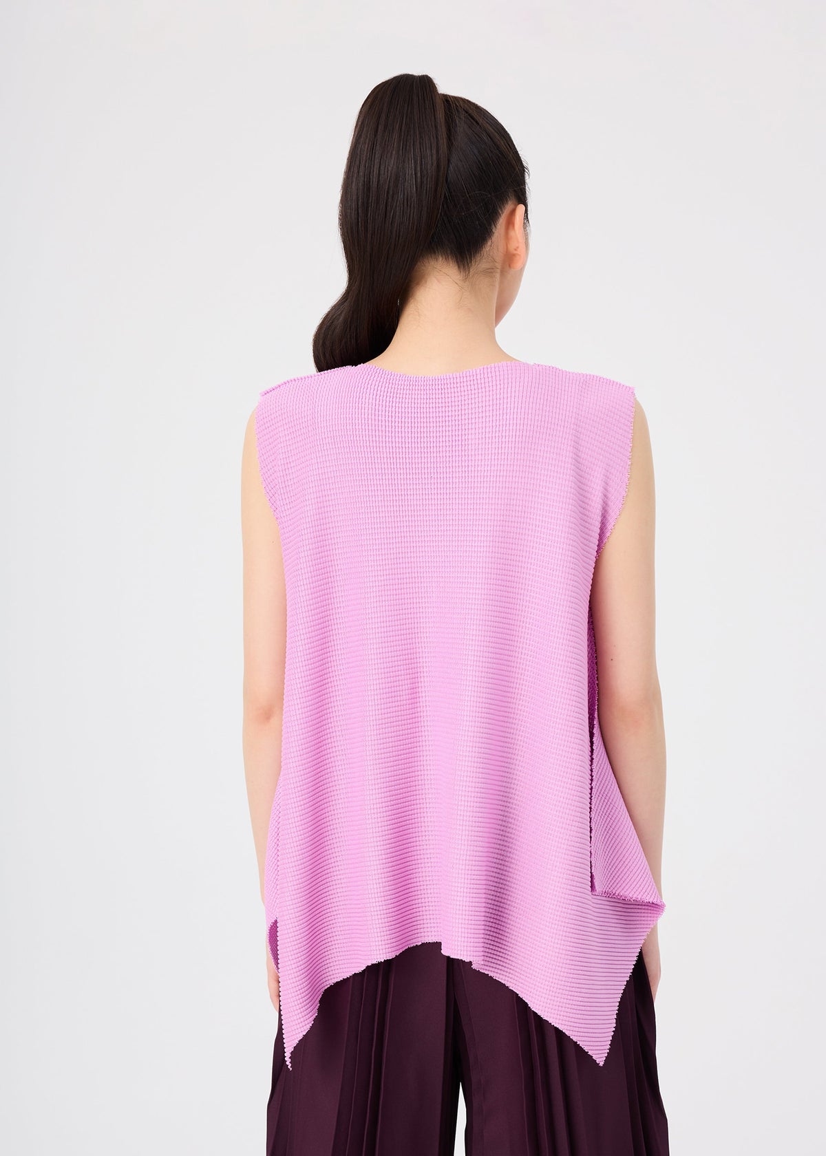 A woman with long dark hair in a ponytail is seen from the back, wearing the TOWER KNIT STRETCH PLEATS sleeveless top by me ISSEY MIYAKE, crafted from soft-textured knit with subtle stretch pleats and featuring an asymmetrical hem. She pairs it with loose dark pants against a plain light background. view 8 of 9