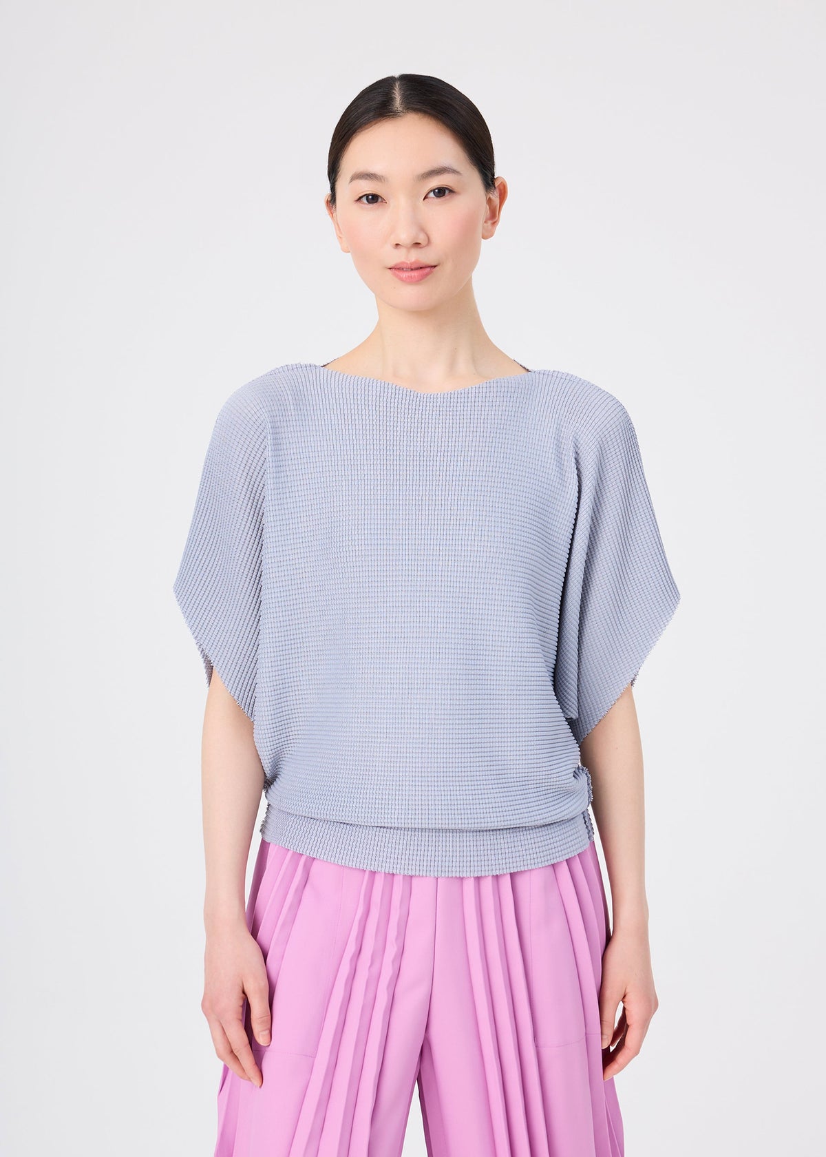 A person with straight dark hair stands facing forward against a plain white background with a neutral expression, wearing a soft-textured, light blue short-sleeved knit top and pink TOWER KNIT STRETCH PLEATS pants by me ISSEY MIYAKE. view 6 of 8