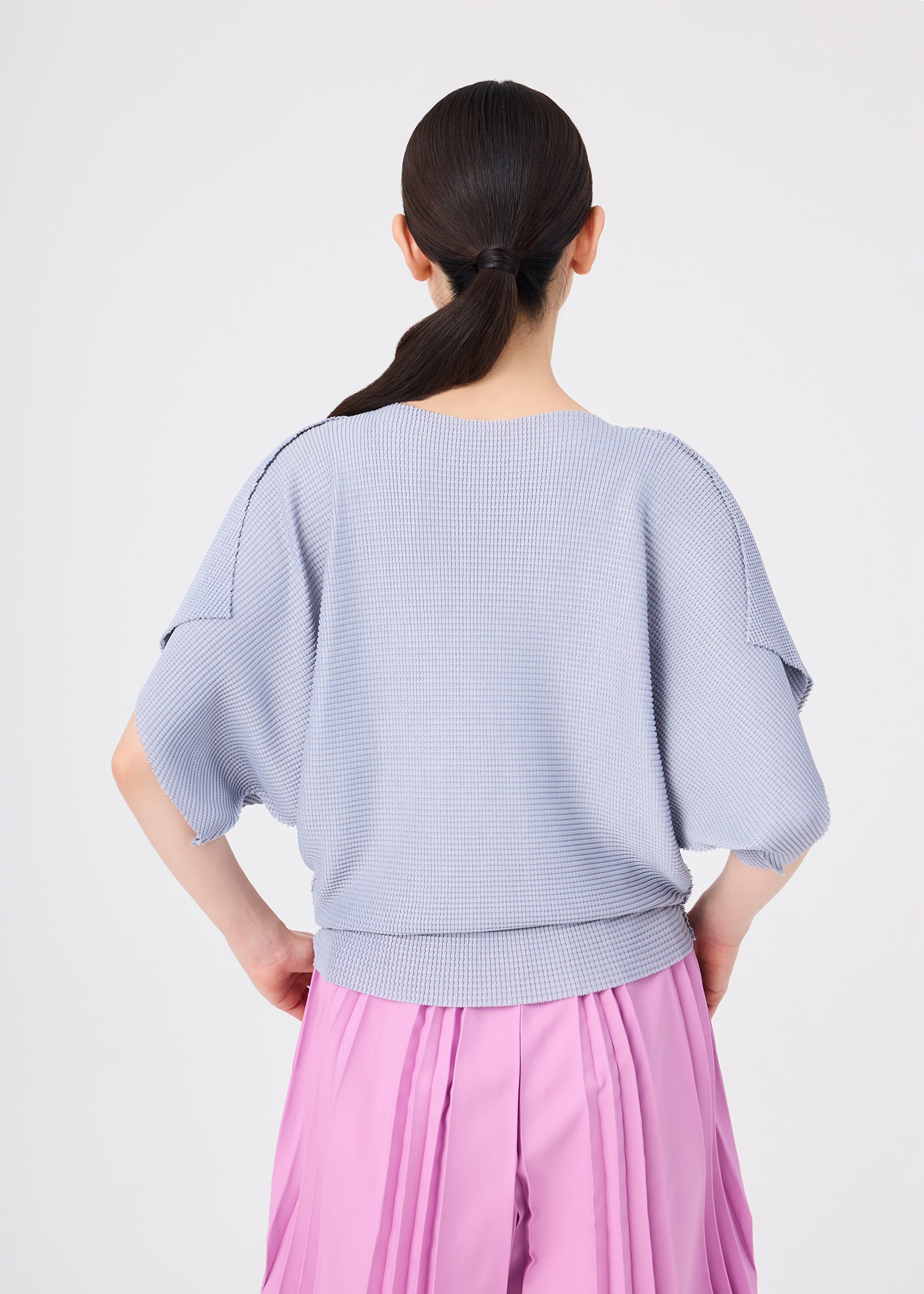 Zoomed view A person with dark hair in a low ponytail is shown from the back, wearing a loose, light purple soft-textured knit short-sleeved top and the bright pink TOWER KNIT STRETCH PLEATS skirt by me ISSEY MIYAKE, featuring knit stretch pleats, against a plain light background.