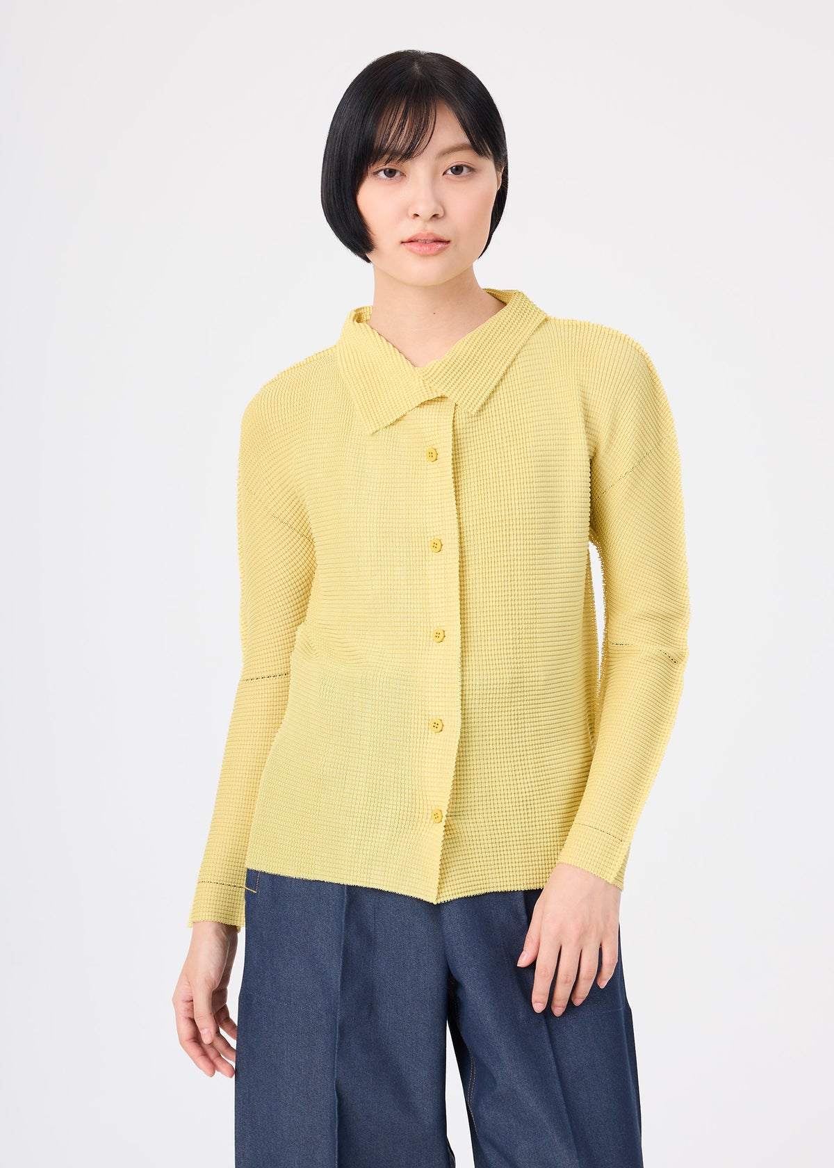 A person stands against a white background wearing me ISSEY MIYAKE KNIT STRETCH PLEATS 1 dark blue pants and a soft, light yellow button-up shirt with a wide collar. view 6 of 8
