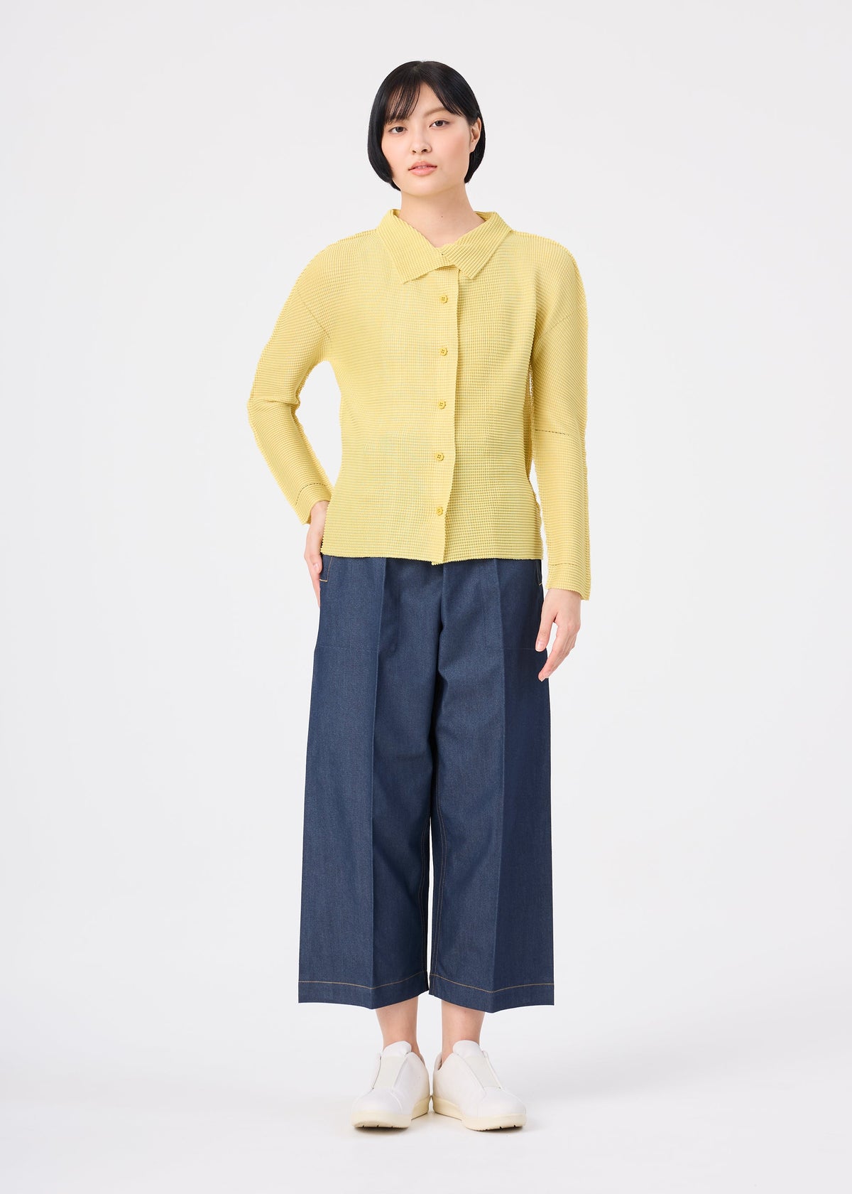 Zoomed view A person stands against a white background wearing me ISSEY MIYAKE KNIT STRETCH PLEATS 1 wide-leg blue cropped pants, a yellow knit cardigan, and white shoes.