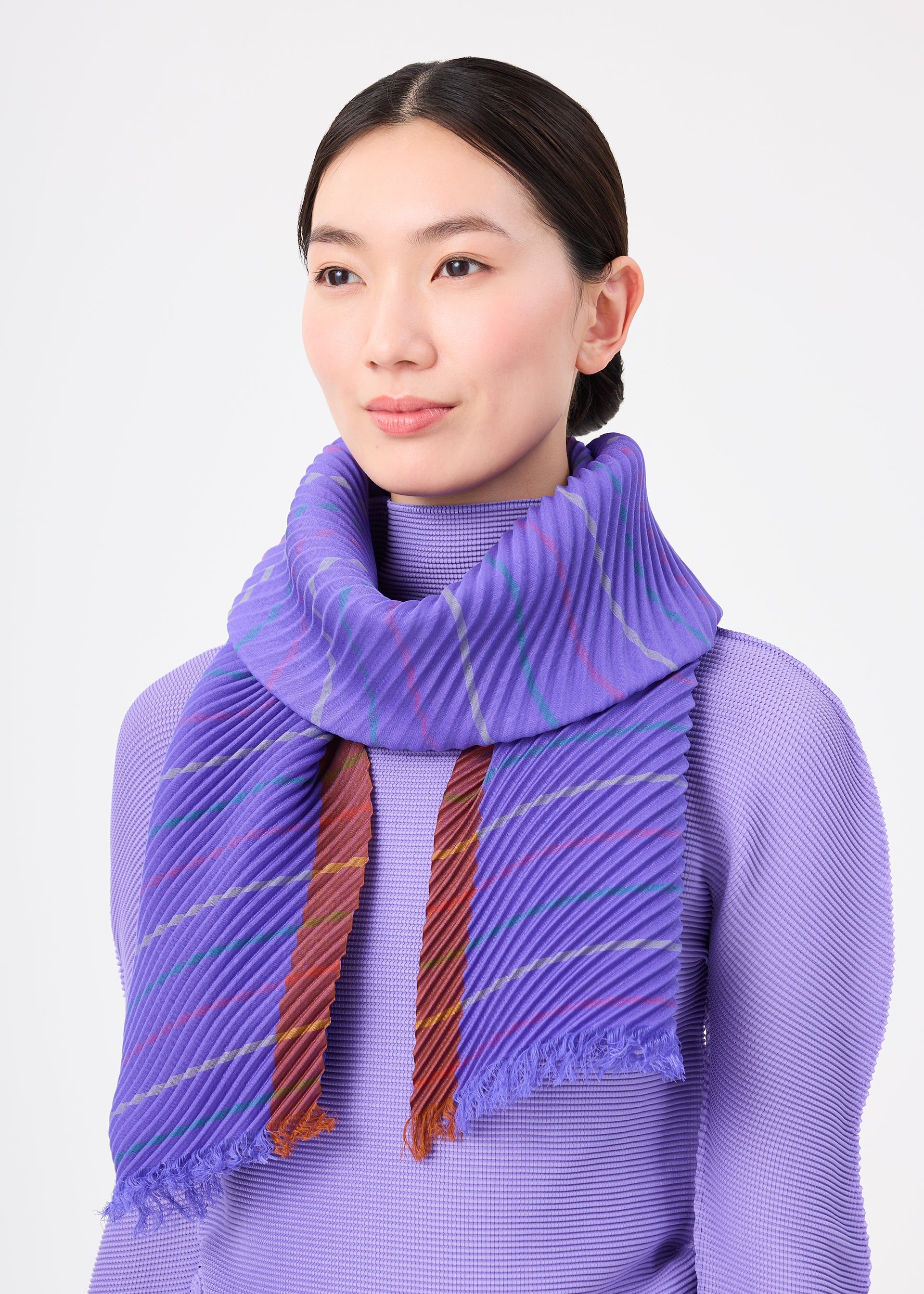Zoomed view A woman with straight dark hair wears a lavender textured top and the STRIPE PLEATS STOLE by me ISSEY MIYAKE, a pleated purple stole with red and blue accents, standing against a plain light background.