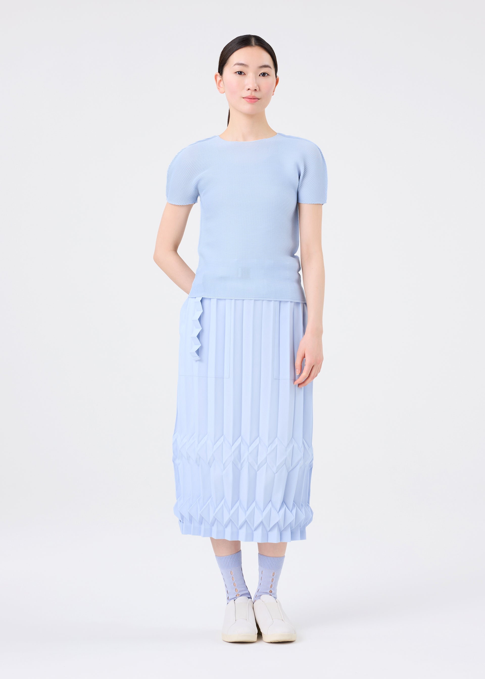 Zoomed view A woman stands against a white background, wearing a light blue short-sleeve top, a matching pleated geometric midi skirt, and me ISSEY MIYAKE’s FRUITS SOCKS with a fun design, paired with white shoes.