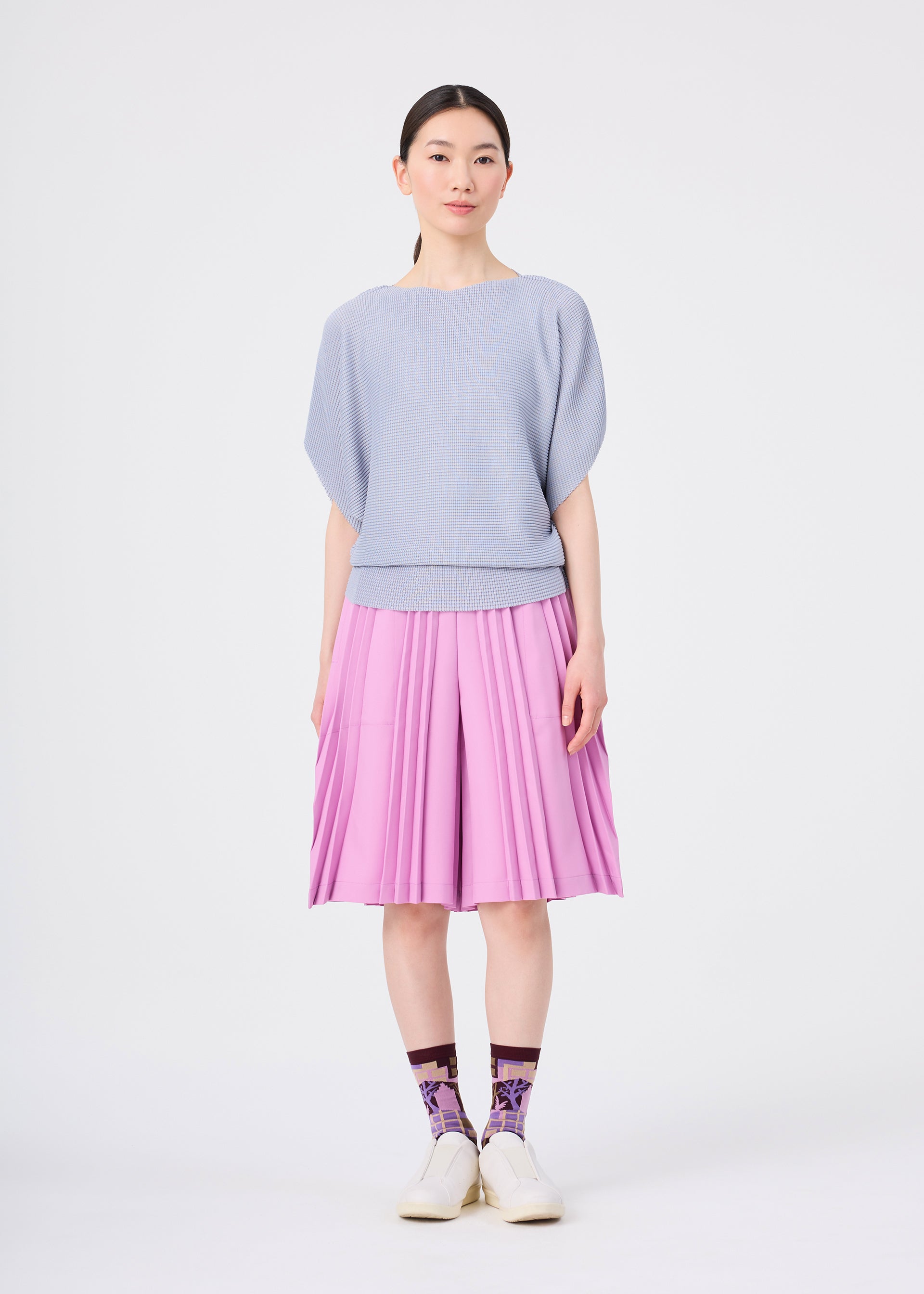 Zoomed view A woman with straight black hair and a neutral expression stands against a plain white background, wearing a light blue textured top, a pink pleated skirt, and striking BUILDING SOCKS by me ISSEY MIYAKE paired with white shoes.