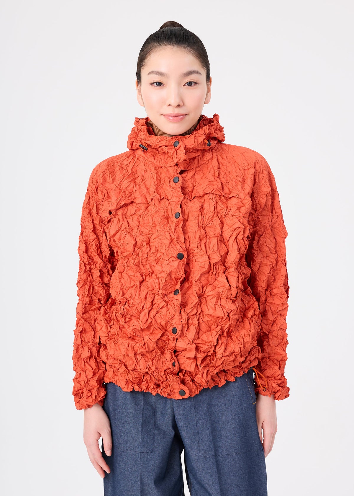 A person stands facing forward against a plain white background, wearing the textured orange MERINGUE PARKA by me ISSEY MIYAKE—a lightweight, water-repellant jacket with a high collar—paired with blue trousers. view 9 of 12