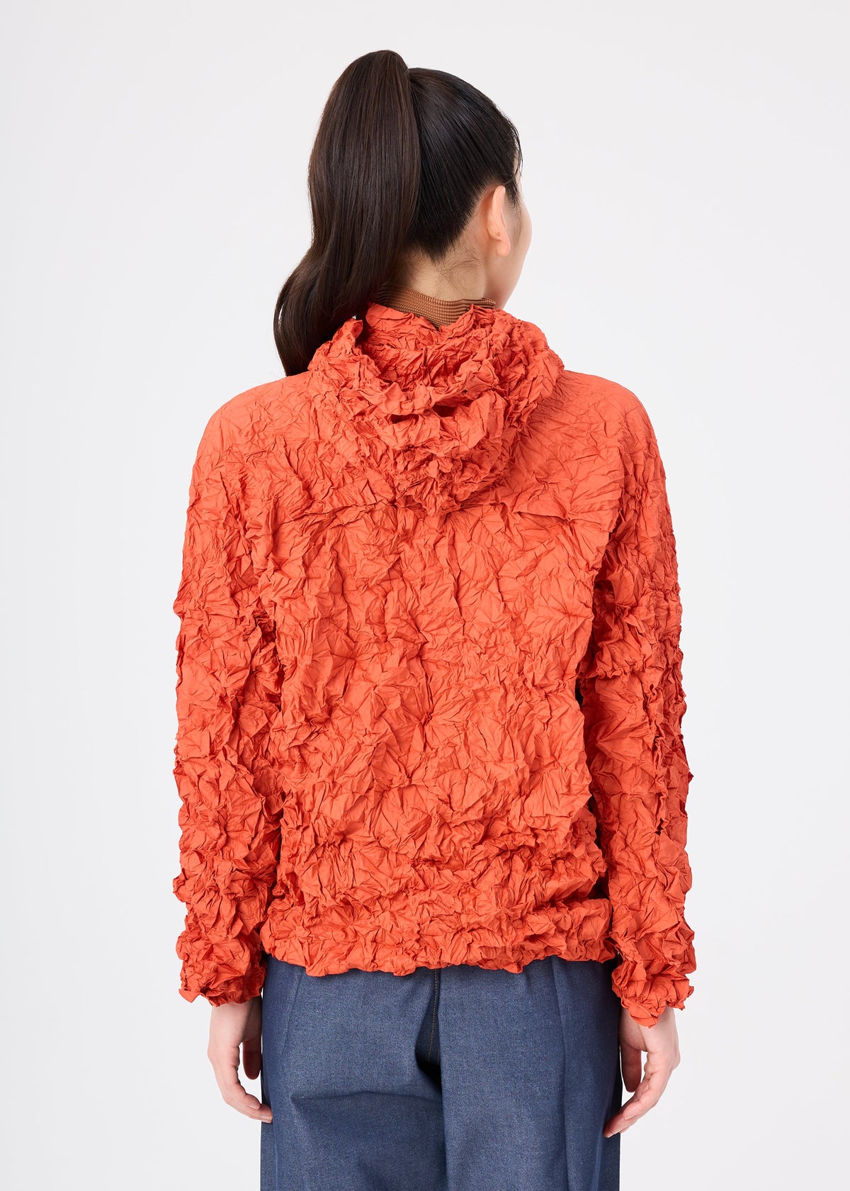 A person with long dark hair in a ponytail wears the MERINGUE PARKA by me ISSEY MIYAKE—a textured, crinkled orange recycled polyester jacket—paired with loose blue pants, facing away from the camera against a plain white background. view 10 of 12