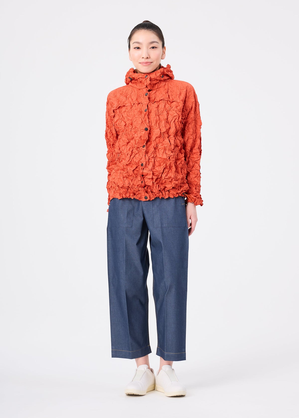 A person stands facing forward against a plain white background, wearing the orange textured lightweight water-repellent MERINGUE PARKA by me ISSEY MIYAKE, paired with blue wide-leg pants and white shoes. view 11 of 12