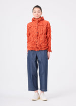 A person stands facing forward against a plain white background, wearing the orange textured lightweight water-repellent MERINGUE PARKA by me ISSEY MIYAKE, paired with blue wide-leg pants and white shoes.