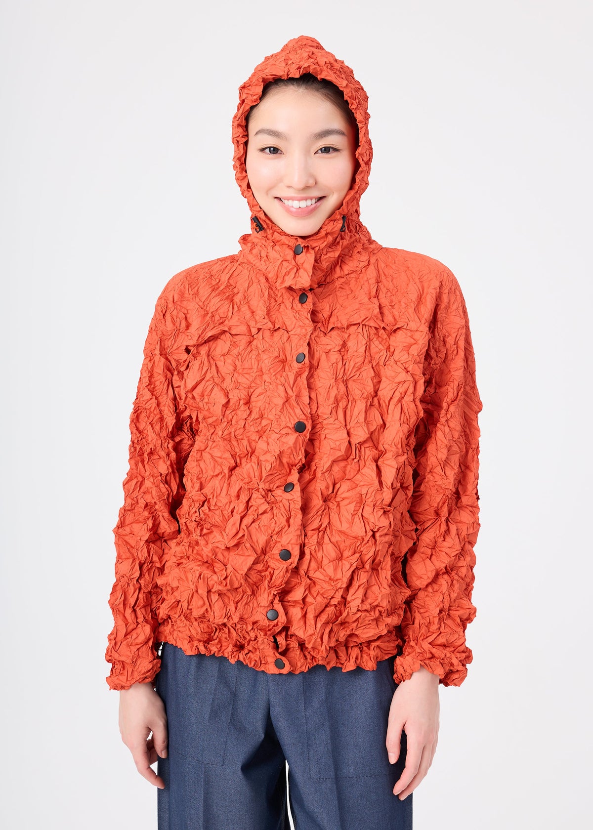 A person smiles while wearing the MERINGUE PARKA by me ISSEY MIYAKE—a textured, crinkled orange packable jacket—paired with blue-gray pants, standing against a plain white background. view 12 of 12
