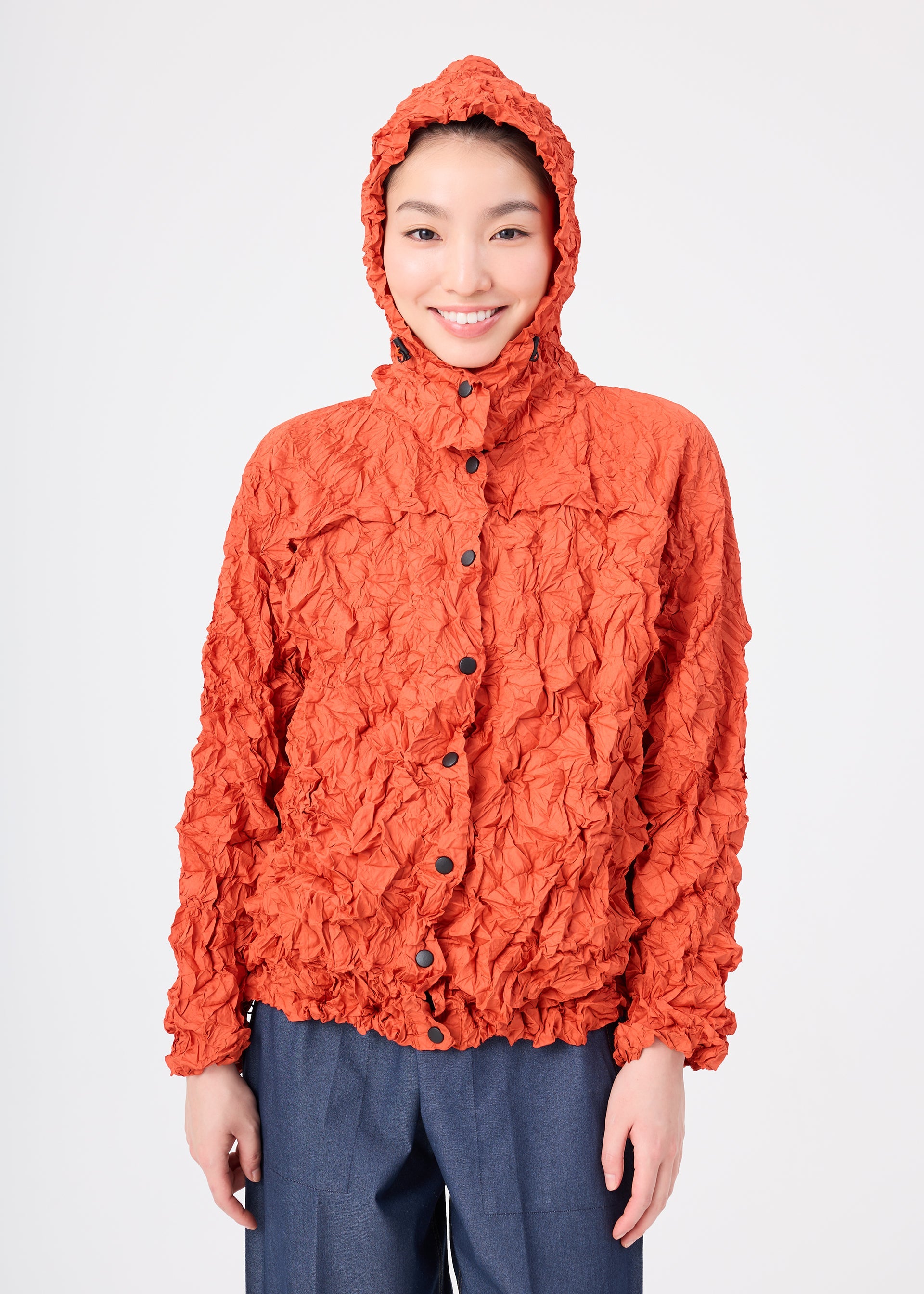 Zoomed view A person smiles while wearing the MERINGUE PARKA by me ISSEY MIYAKE—a textured, crinkled orange packable jacket—paired with blue-gray pants, standing against a plain white background.