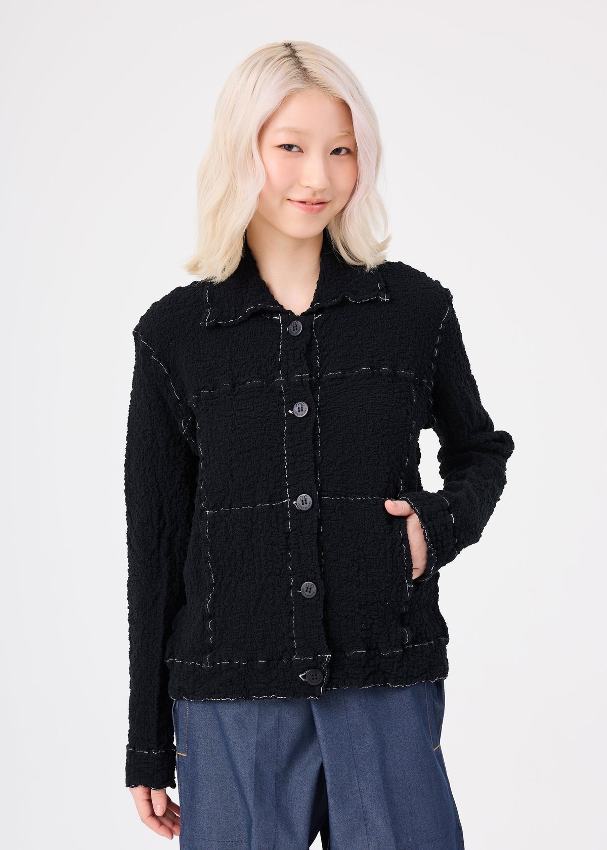 A person with light blonde, wavy hair wears the me ISSEY MIYAKE CAULIFLOWER COLOR 1 black textured button-up jacket and blue pants, standing with one hand in their pocket and smiling against a plain white background. view 6 of 8