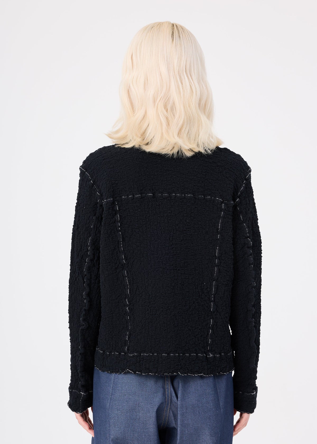 A person with shoulder-length, wavy blonde hair is seen from the back wearing the me ISSEY MIYAKE CAULIFLOWER COLOR 1 black textured jacket, styled with blue pants against a plain white background. view 7 of 8