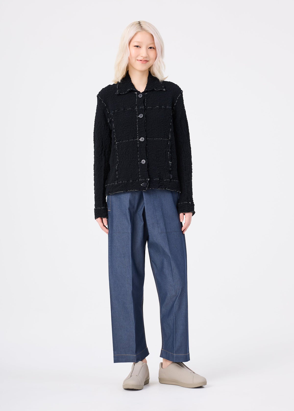 A person with light blonde hair faces forward, wearing the CAULIFLOWER COLOR 1 black textured button-up jacket by me ISSEY MIYAKE, loose blue trousers, and beige slip-on shoes against a plain white background. view 8 of 8