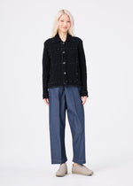 A person with light blonde hair faces forward, wearing the CAULIFLOWER COLOR 1 black textured button-up jacket by me ISSEY MIYAKE, loose blue trousers, and beige slip-on shoes against a plain white background.