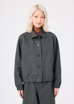A person with light blonde, wavy hair is wearing the me ISSEY MIYAKE PINA CLOTH dark gray, garment-dyed jacket and matching pants, standing against a plain white background, facing forward and smiling slightly.