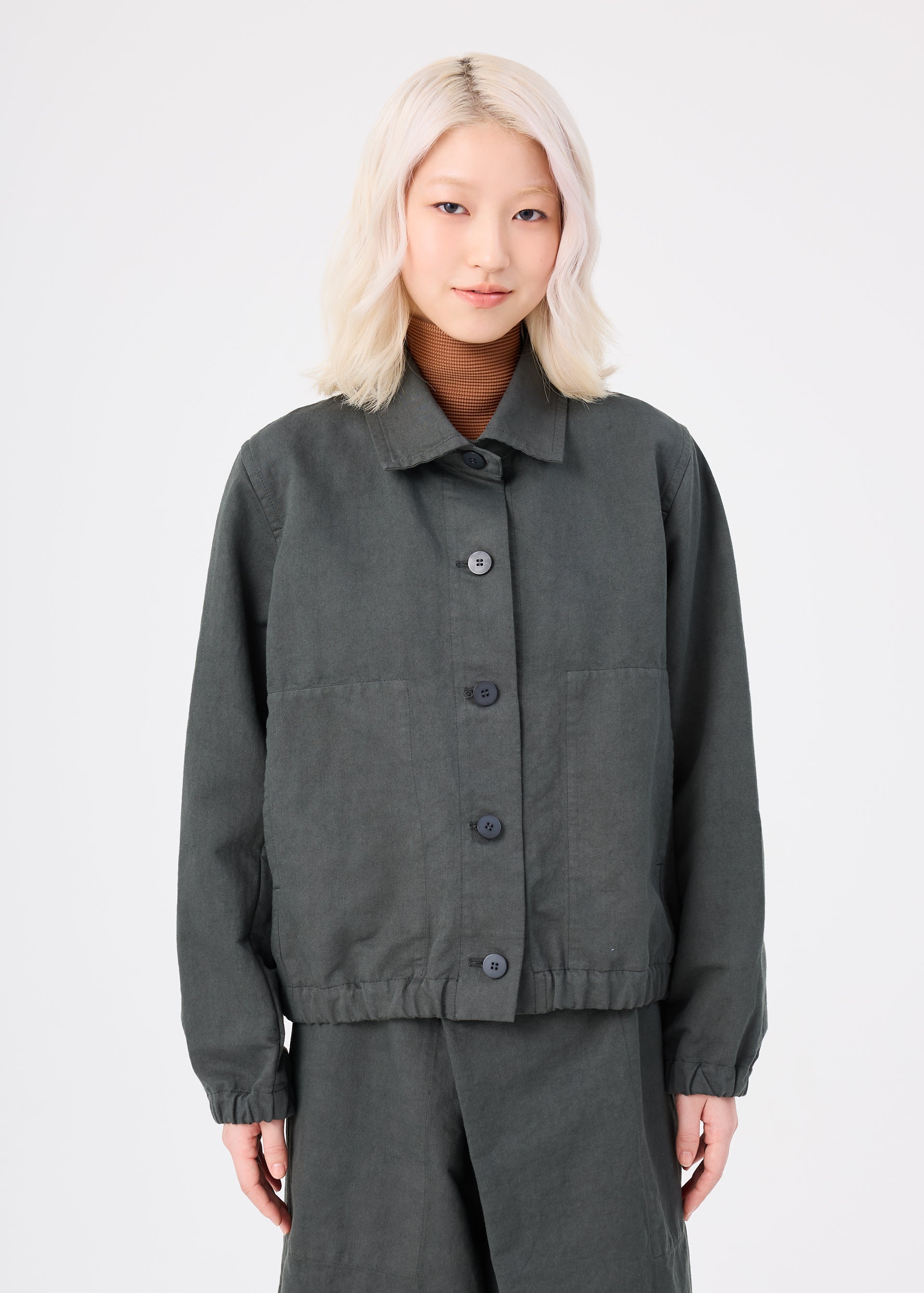Zoomed view A person with light blonde, wavy hair is wearing the me ISSEY MIYAKE PINA CLOTH dark gray, garment-dyed jacket and matching pants, standing against a plain white background, facing forward and smiling slightly.