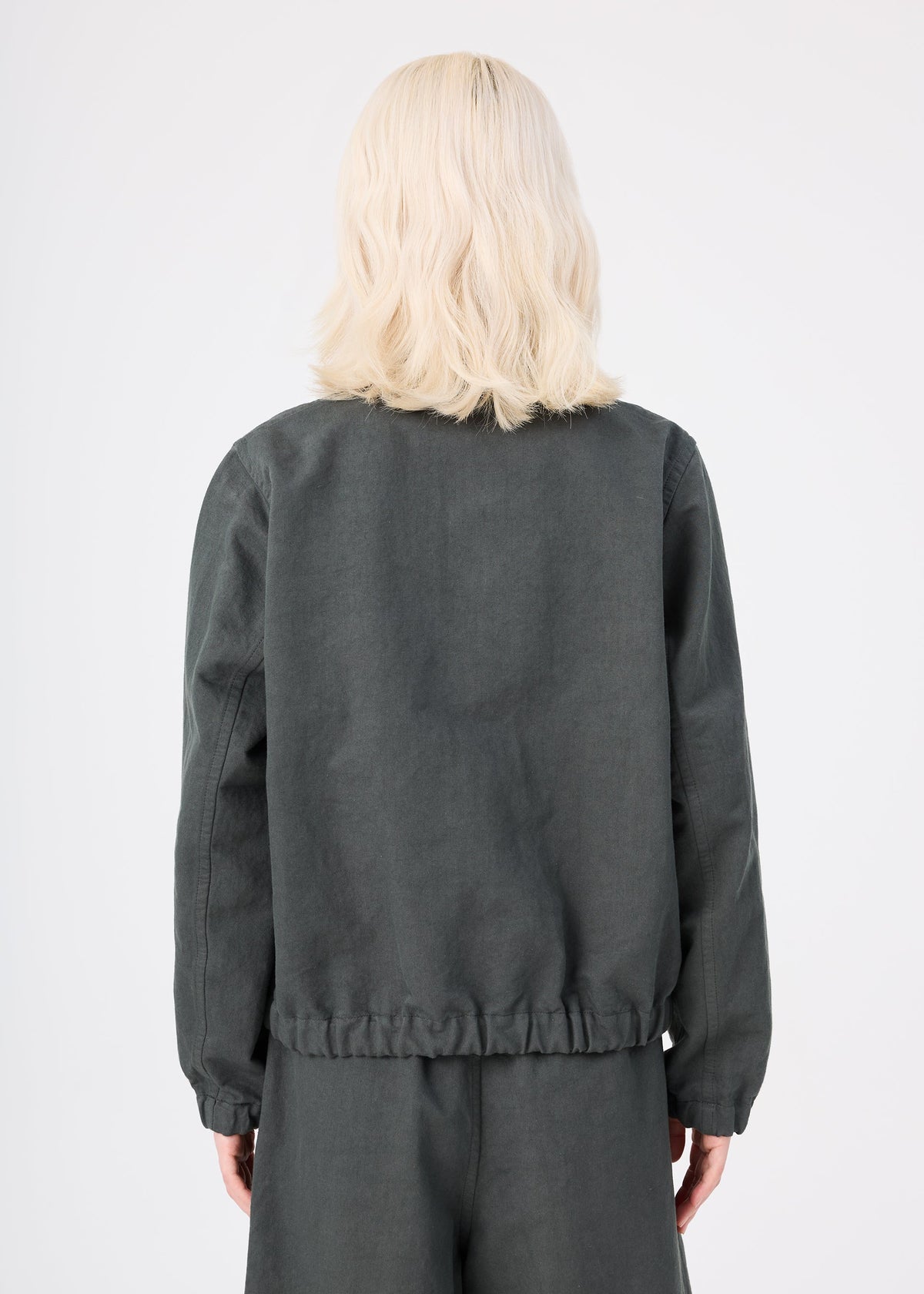 A person with shoulder-length blonde hair, wearing the dark gray PINA CLOTH jacket and matching pants by me ISSEY MIYAKE, stands with their back to the camera against a plain light background. view 6 of 8