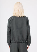 A person with shoulder-length blonde hair, wearing the dark gray PINA CLOTH jacket and matching pants by me ISSEY MIYAKE, stands with their back to the camera against a plain light background.