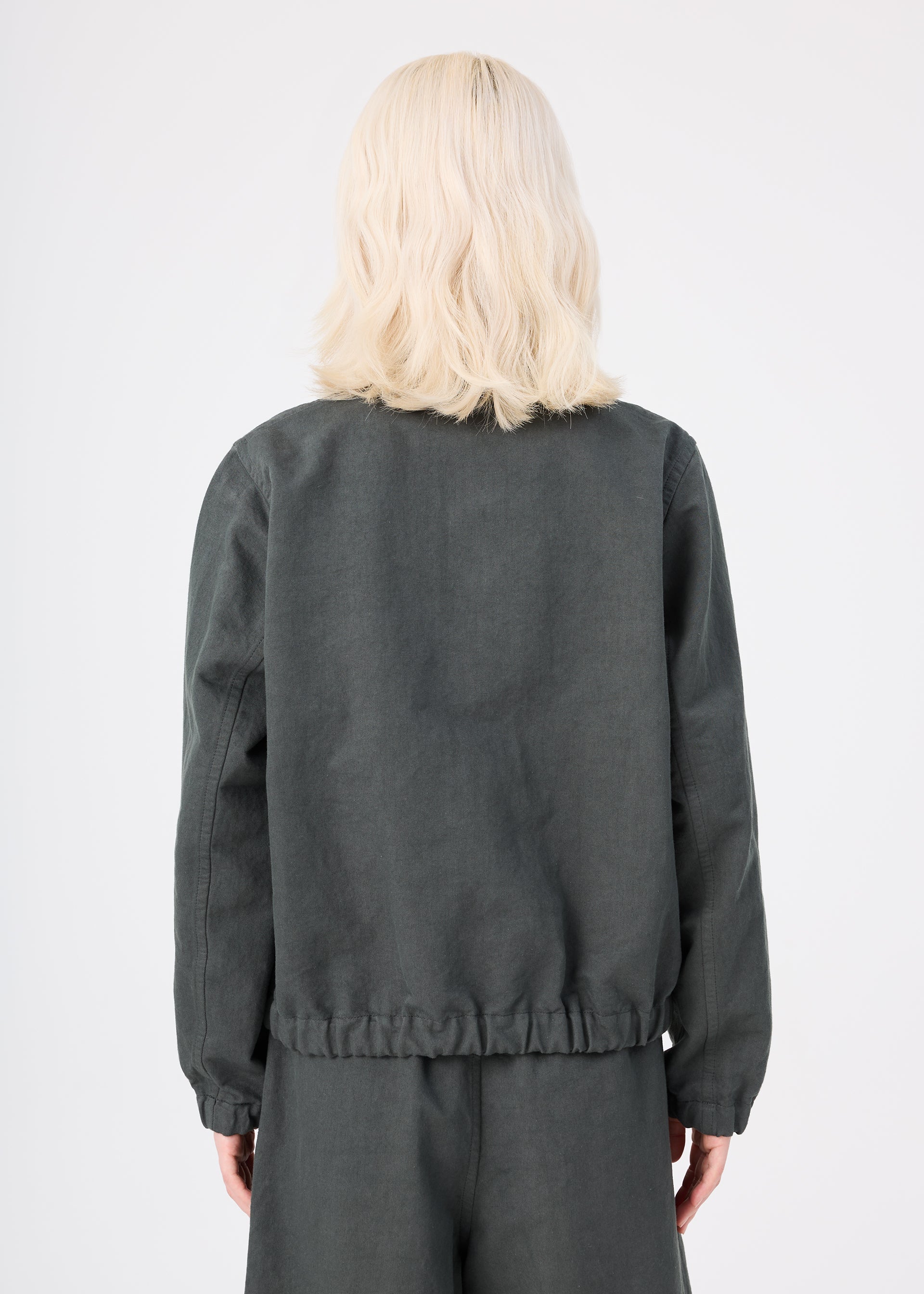 Zoomed view A person with shoulder-length blonde hair, wearing the dark gray PINA CLOTH jacket and matching pants by me ISSEY MIYAKE, stands with their back to the camera against a plain light background.