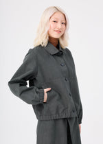 A person with light blonde hair is wearing the me ISSEY MIYAKE PINA CLOTH garment-dyed jacket and matching pants, standing against a plain white background and smiling gently with one hand in a pocket.
